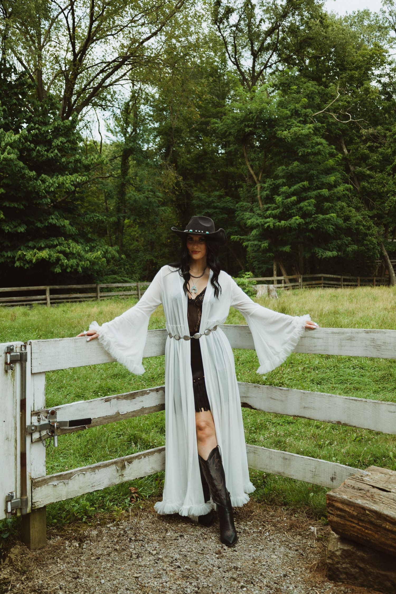 Person in flowing white dress and cowboy hat poses near wooden fence in a lush green field.