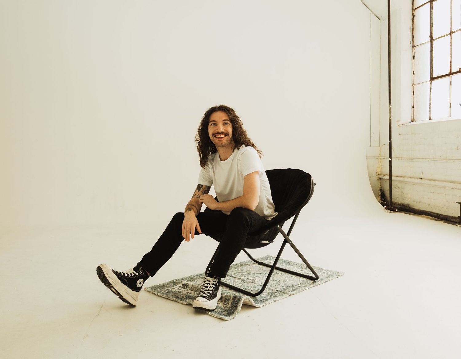 A person with long dark hair and a mustache sits casually on a folding chair in a bright white room.