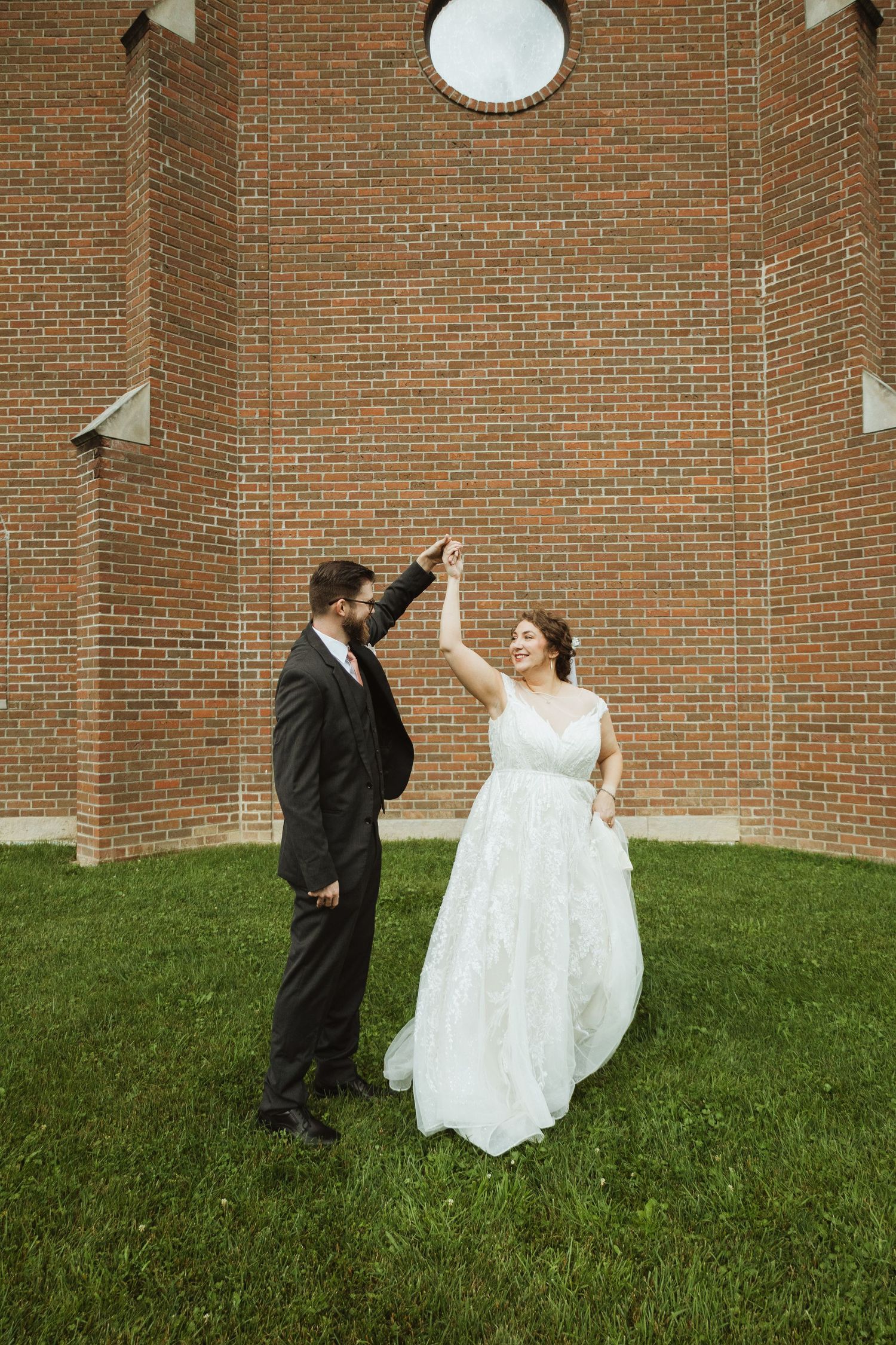 A playful wedding photoshoot on a grassy lawn against a brick church wall.