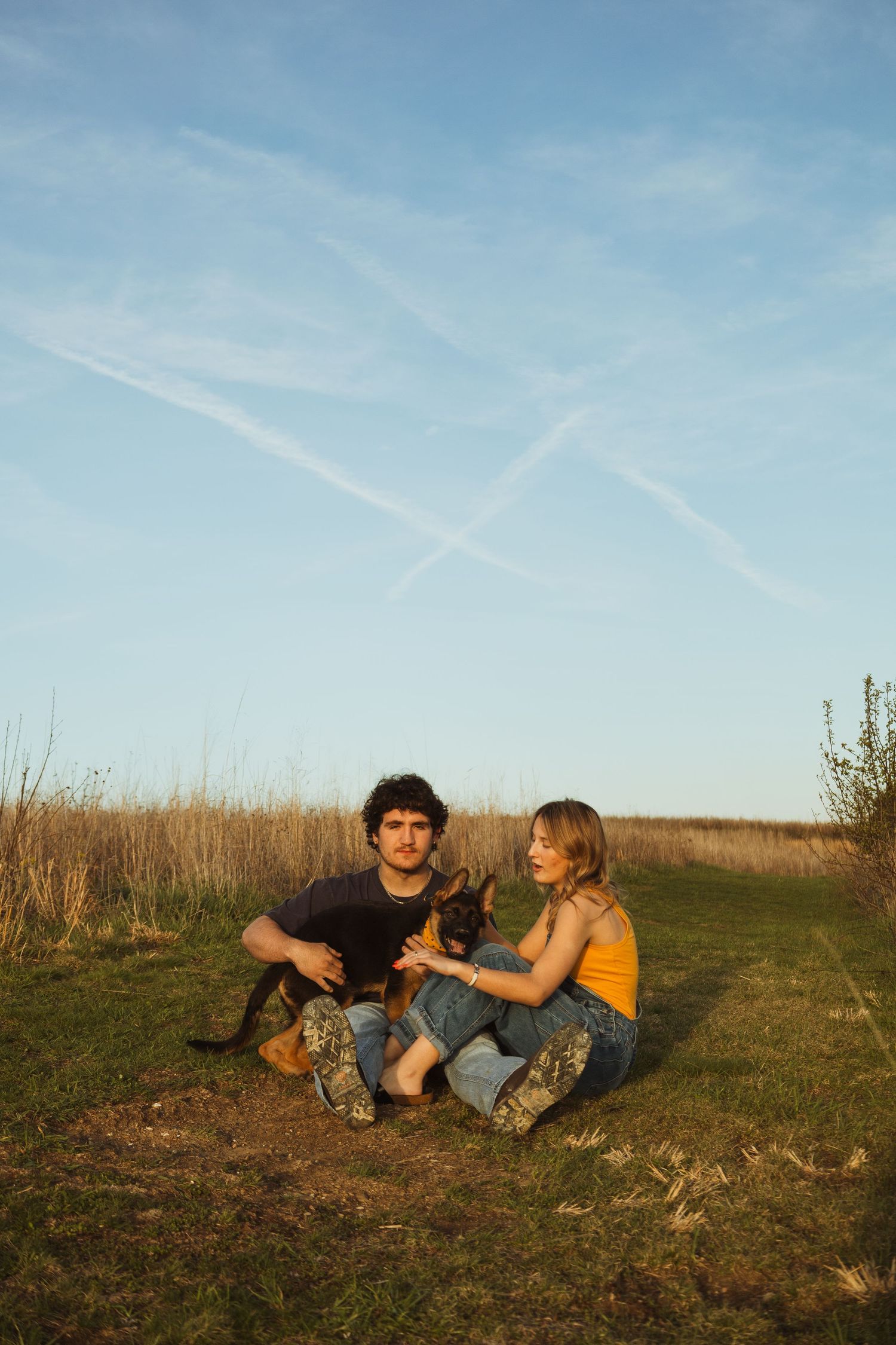 A young couple sits on grass in a field, petting a small dog under a blue sky with contrail patterns.