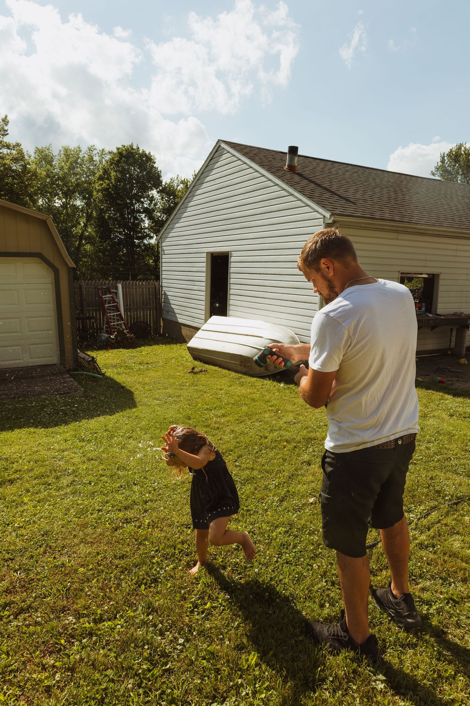 A father plays with his toddler in a sunny backyard, holding a toy while the child runs on green grass near a white house.