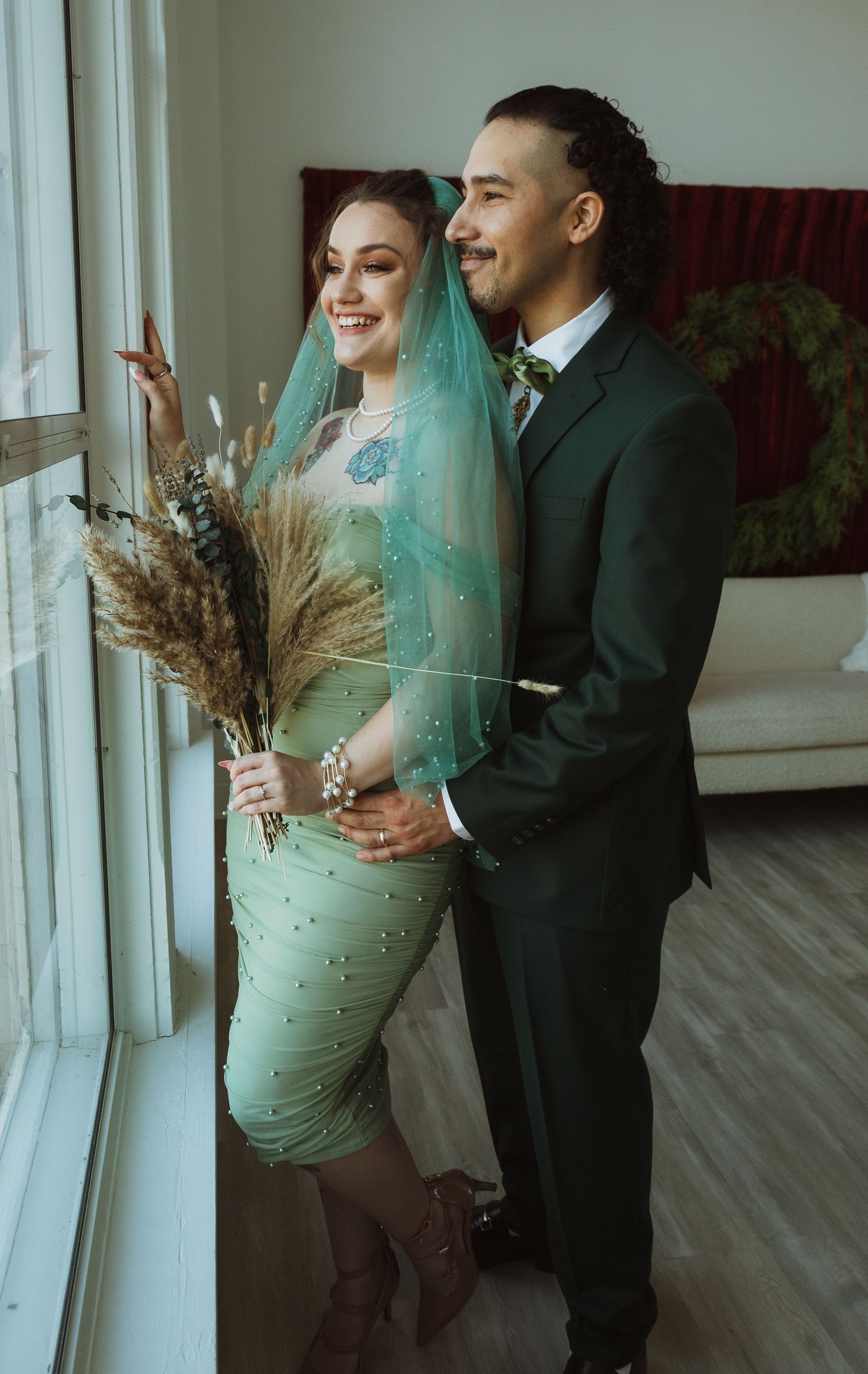Smiling newlyweds in green wedding attire looking out a bright window together.