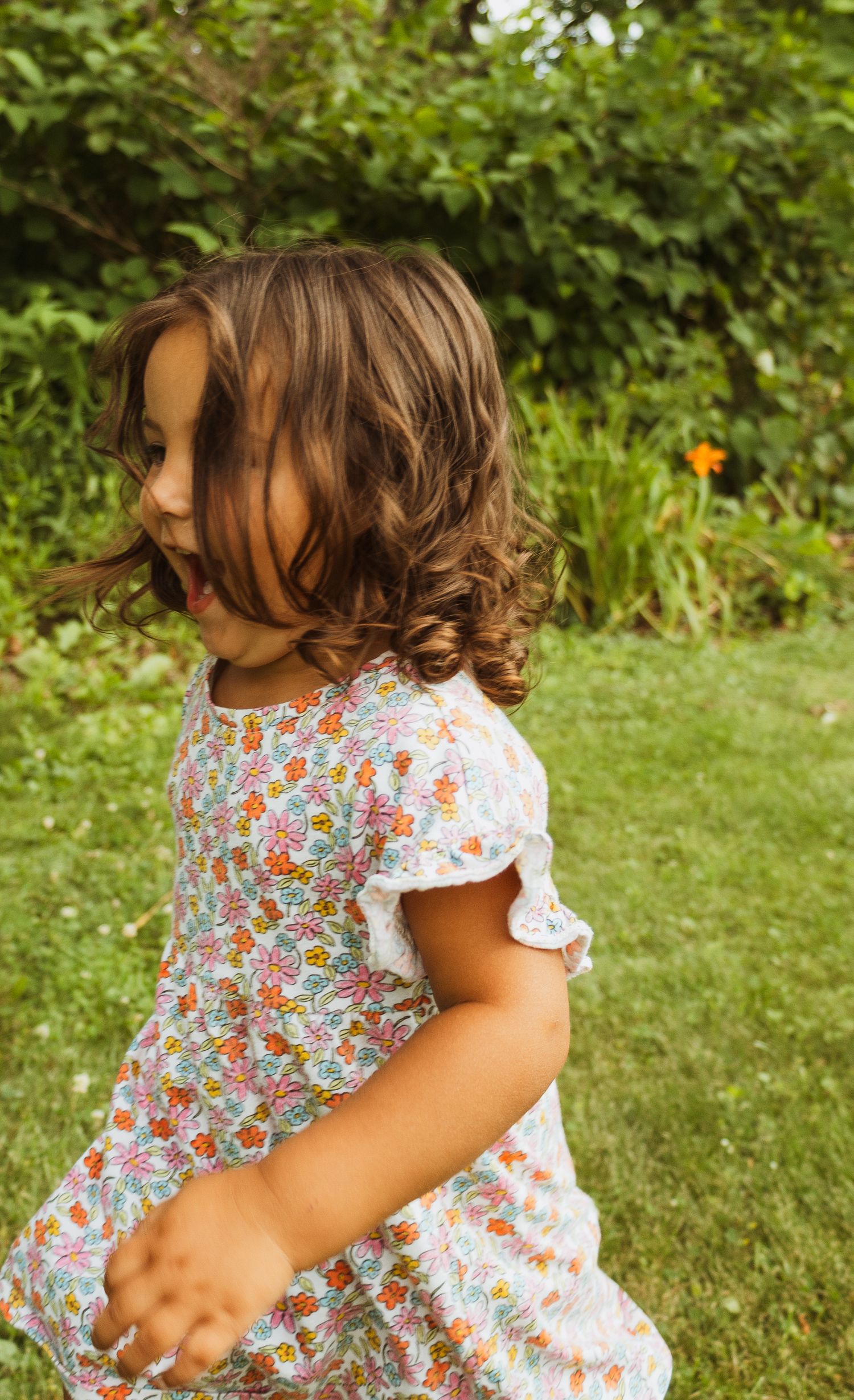 A young girl with curly brown hair twirls in a floral dress on a green lawn surrounded by lush garden foliage.