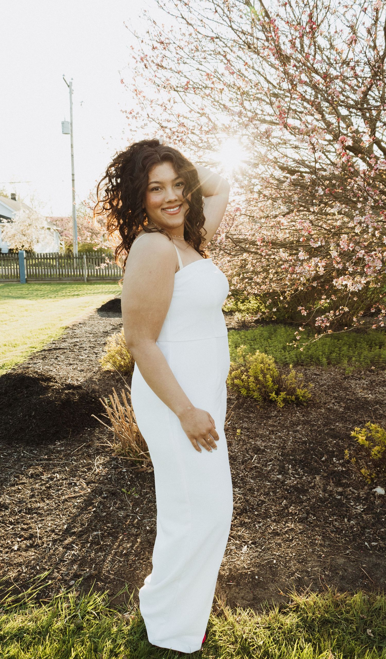 A woman in white poses gracefully by blooming spring trees at sunset.