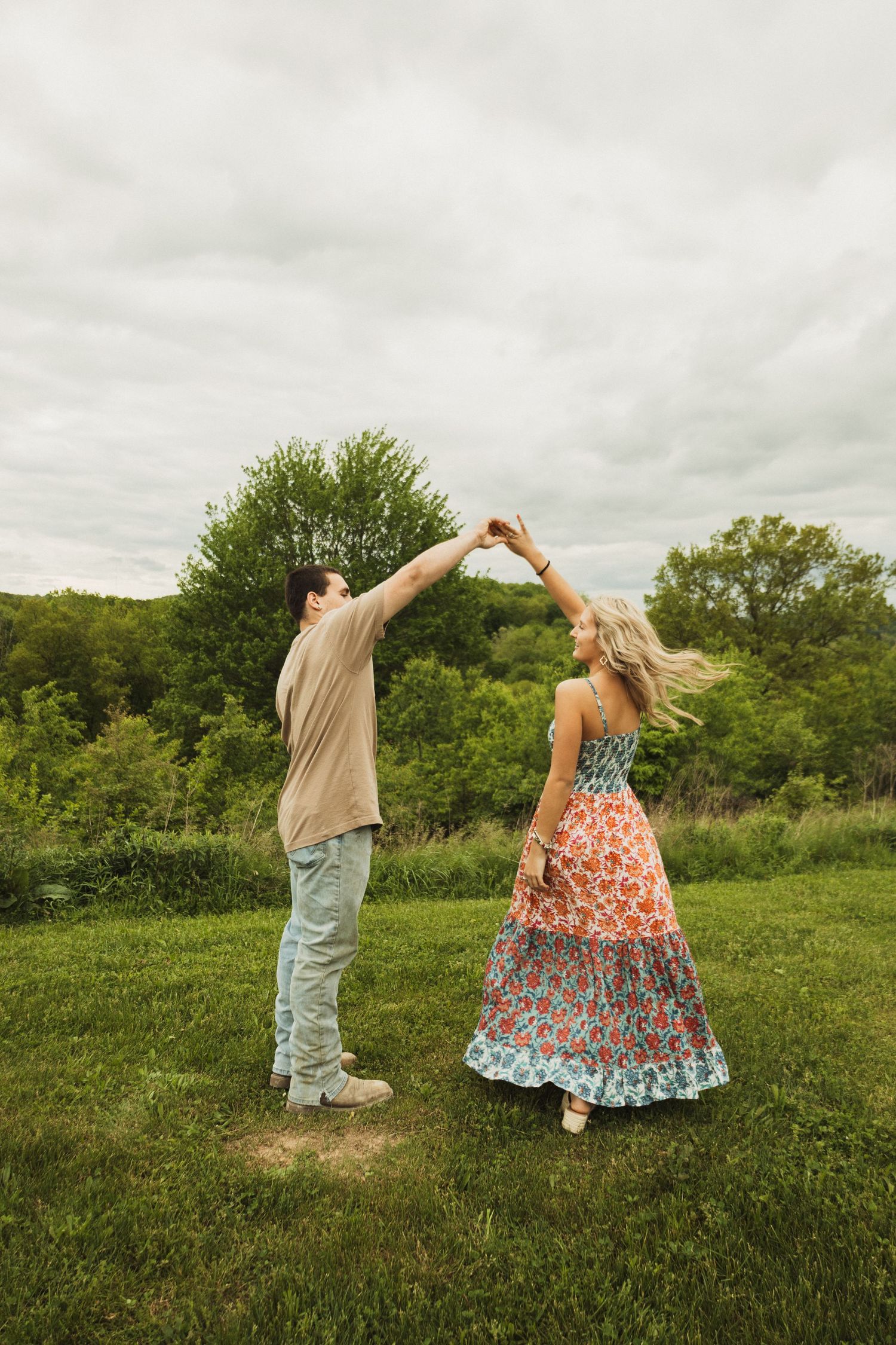 A couple shares a playful dance moment in a scenic meadow surrounded by lush greenery.