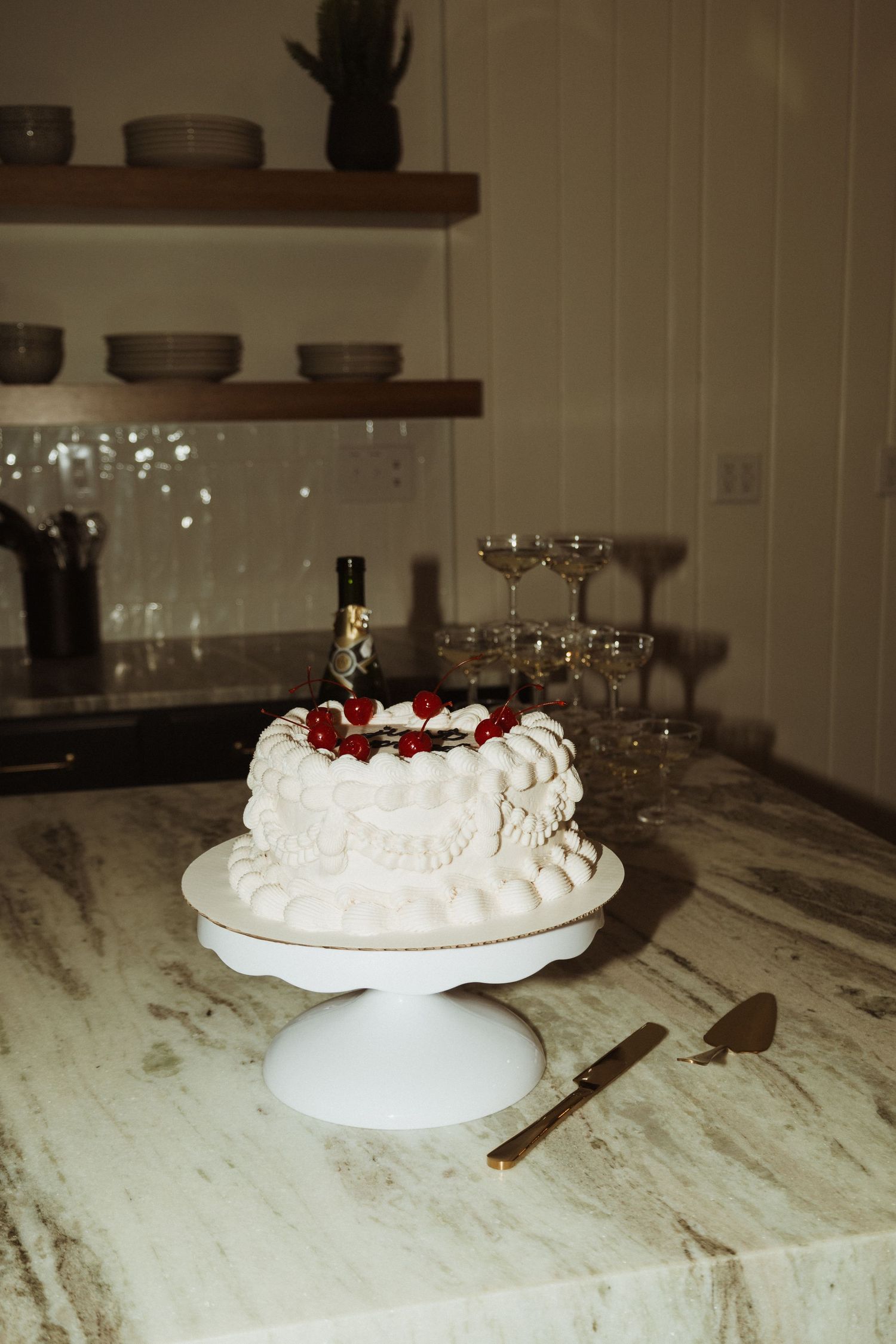Wedding cake display on white pedestal with champagne glasses and rustic wooden shelving in background.