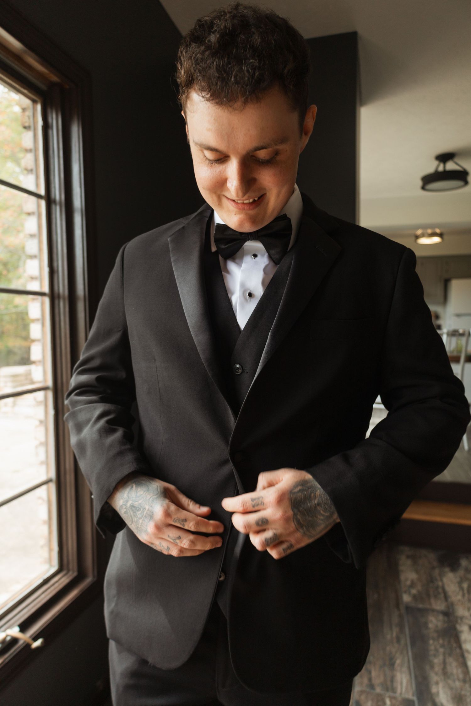A groom in a classic black tuxedo with bow tie buttons his jacket while standing by a window.