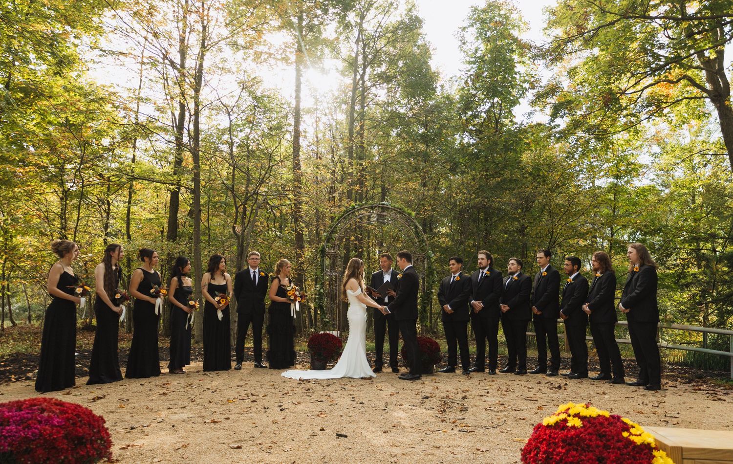 Wedding ceremony group photo with bridal party arranged in line among autumn trees.