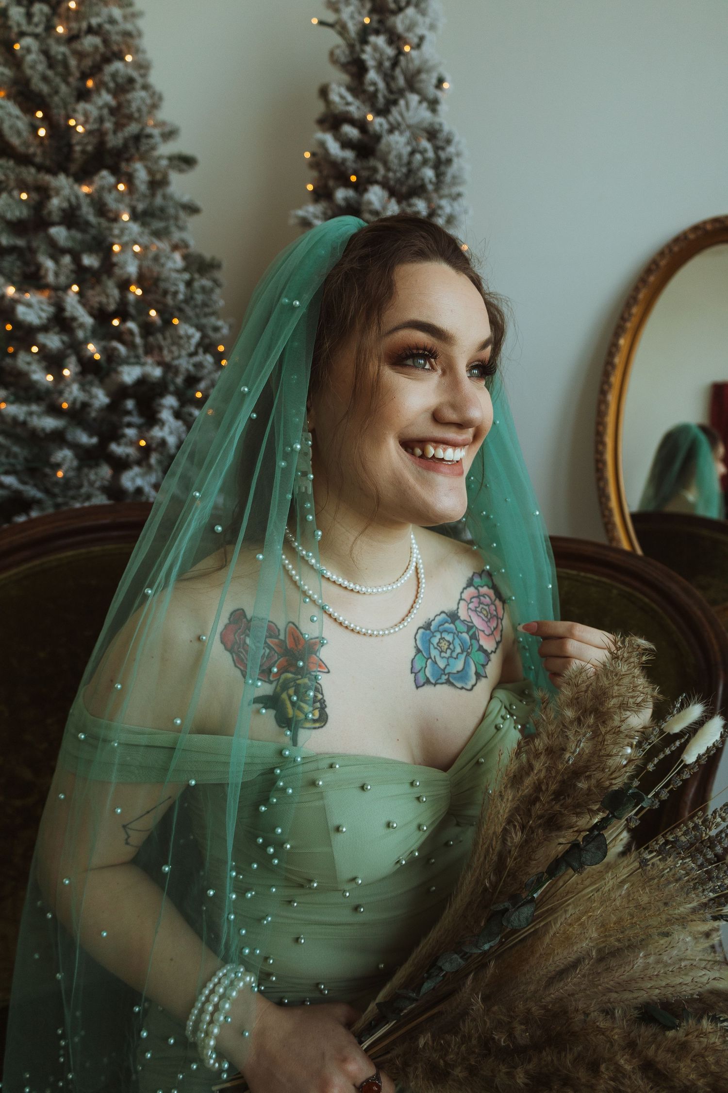 Tattooed bride in green gown and teal pearl veil laughing beside Christmas trees holding dried bouquet.