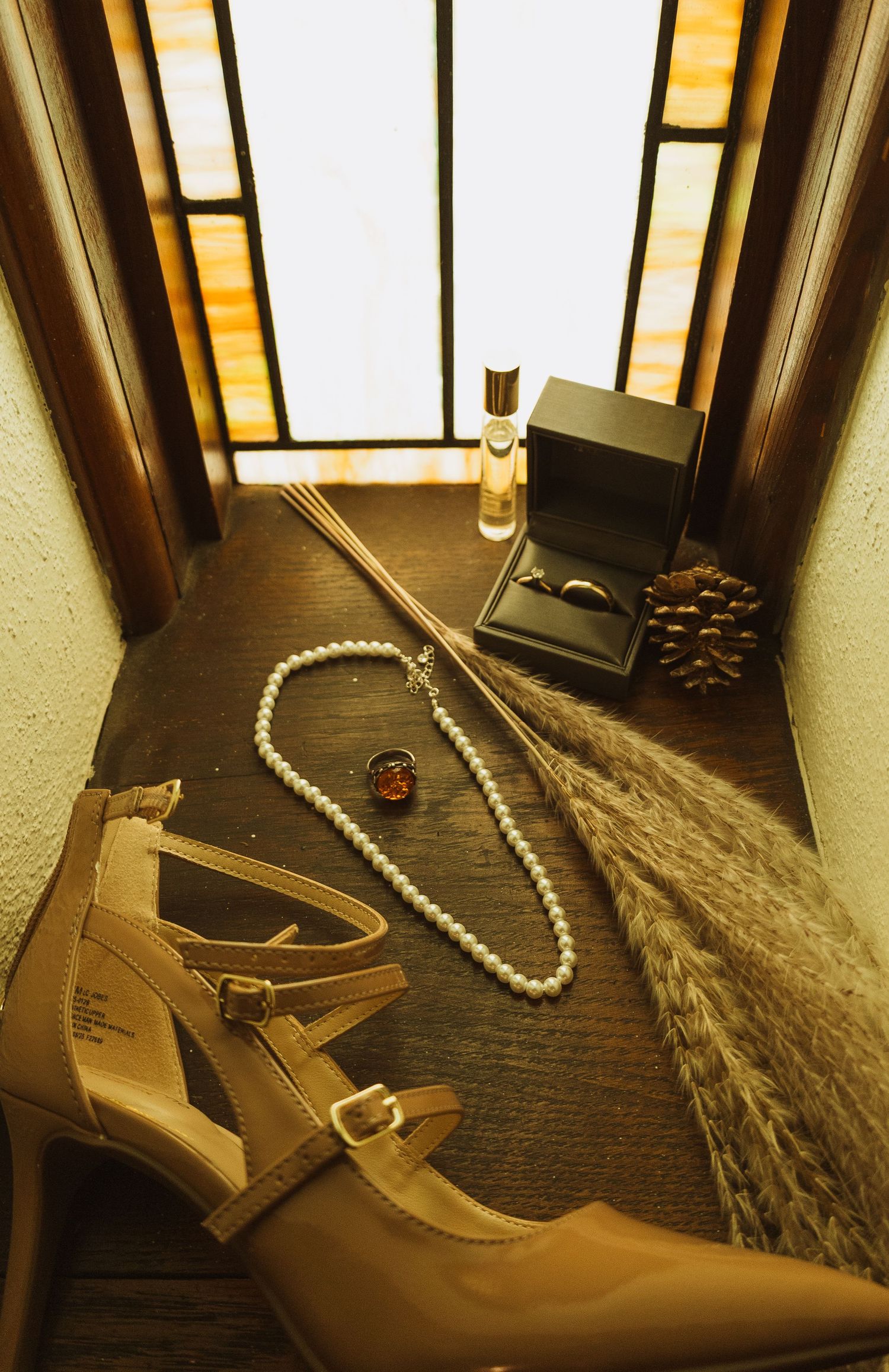 Bridal flat lay with pearl necklace, heels, ring box, perfume, and dried pampas grass on wooden surface near window.