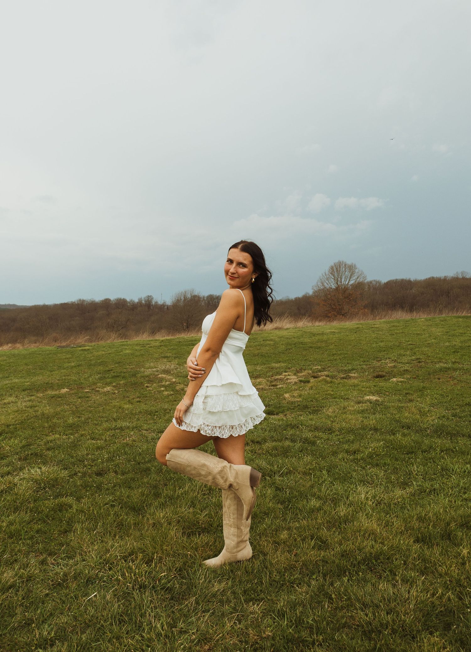 Woman in white mini dress and tan cowboy boots posing in a green field under a cloudy sky.
