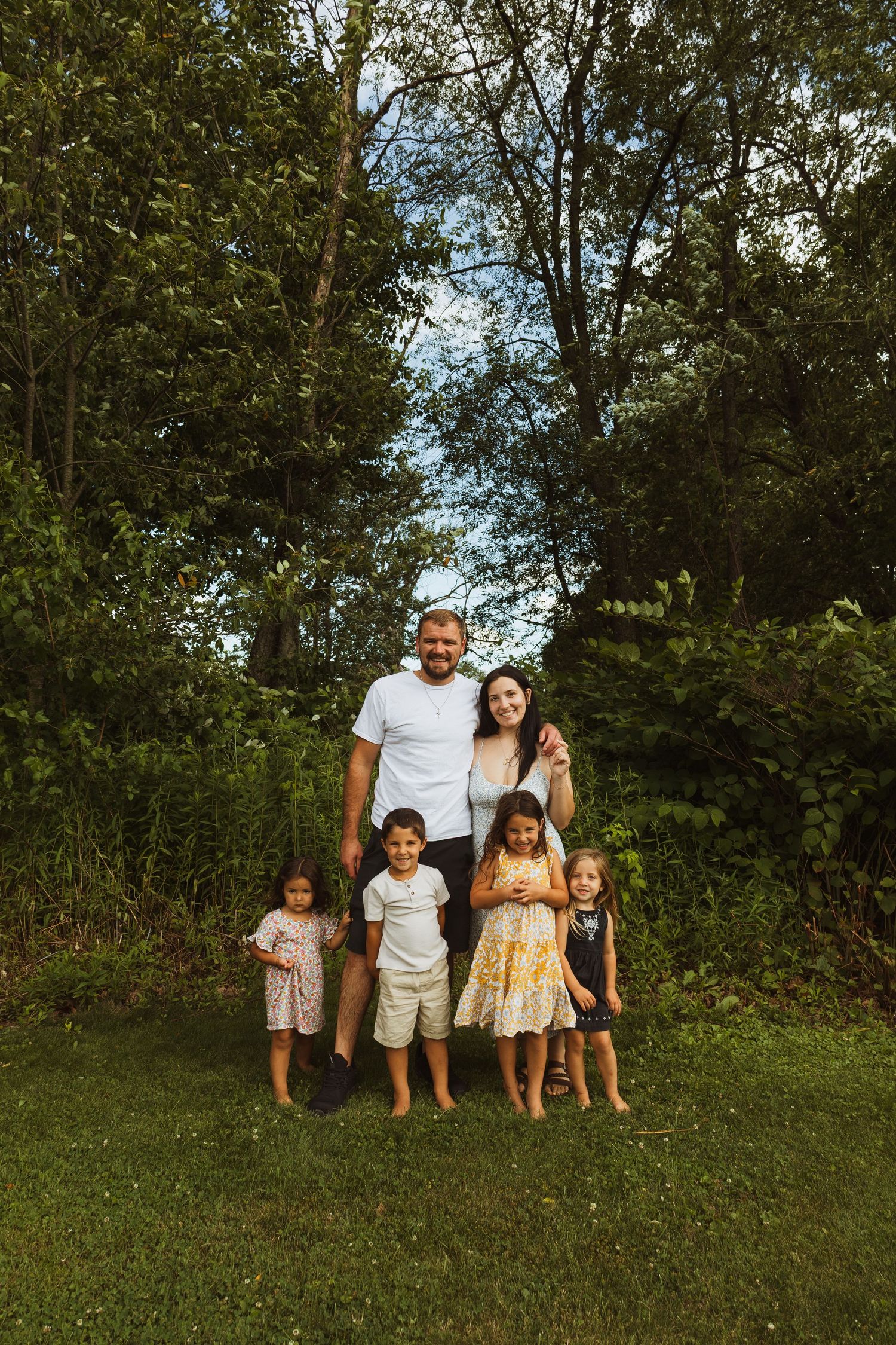 Family of six posing outdoors on green grass surrounded by lush trees, parents standing behind four young children.
