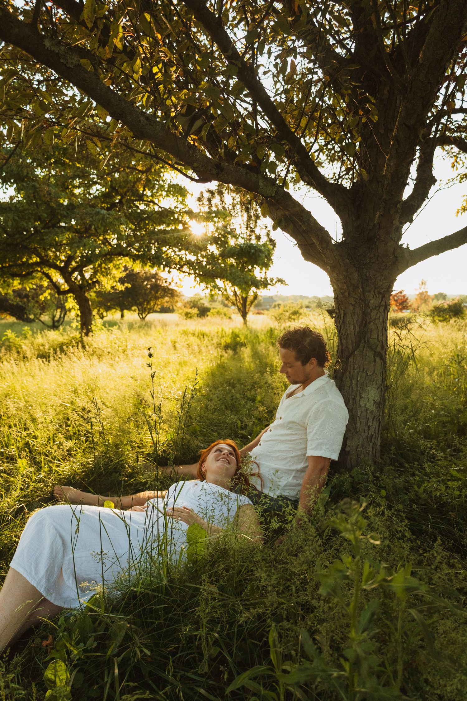 Couple relaxing under a tree in a sunny green meadow, woman lying in grass while man sits against trunk at golden hour.