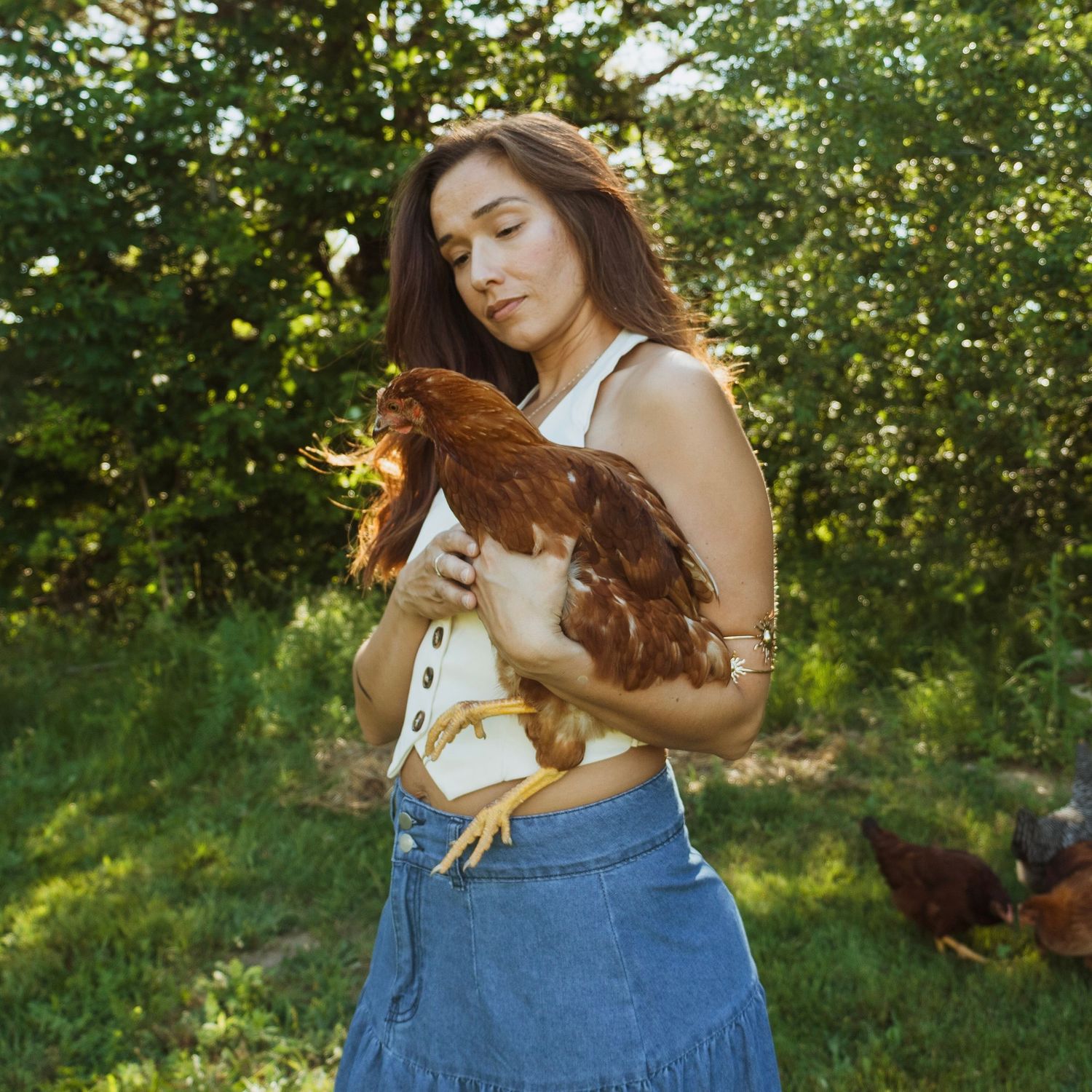 Young woman in white top and denim skirt holds a brown chicken outdoors on a green farm.