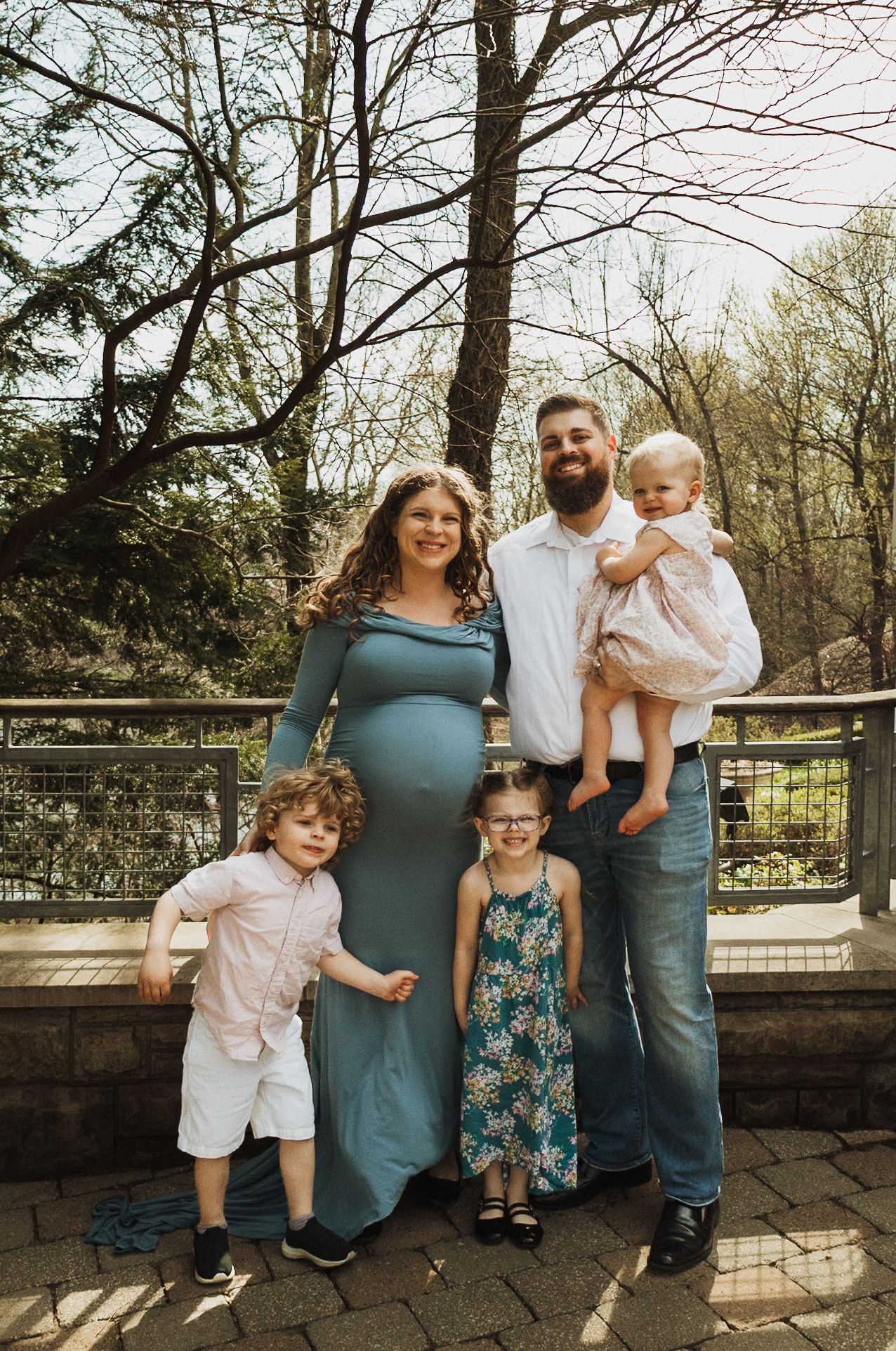 Family of five poses together on an outdoor stone patio surrounded by bare trees on a bright spring day.