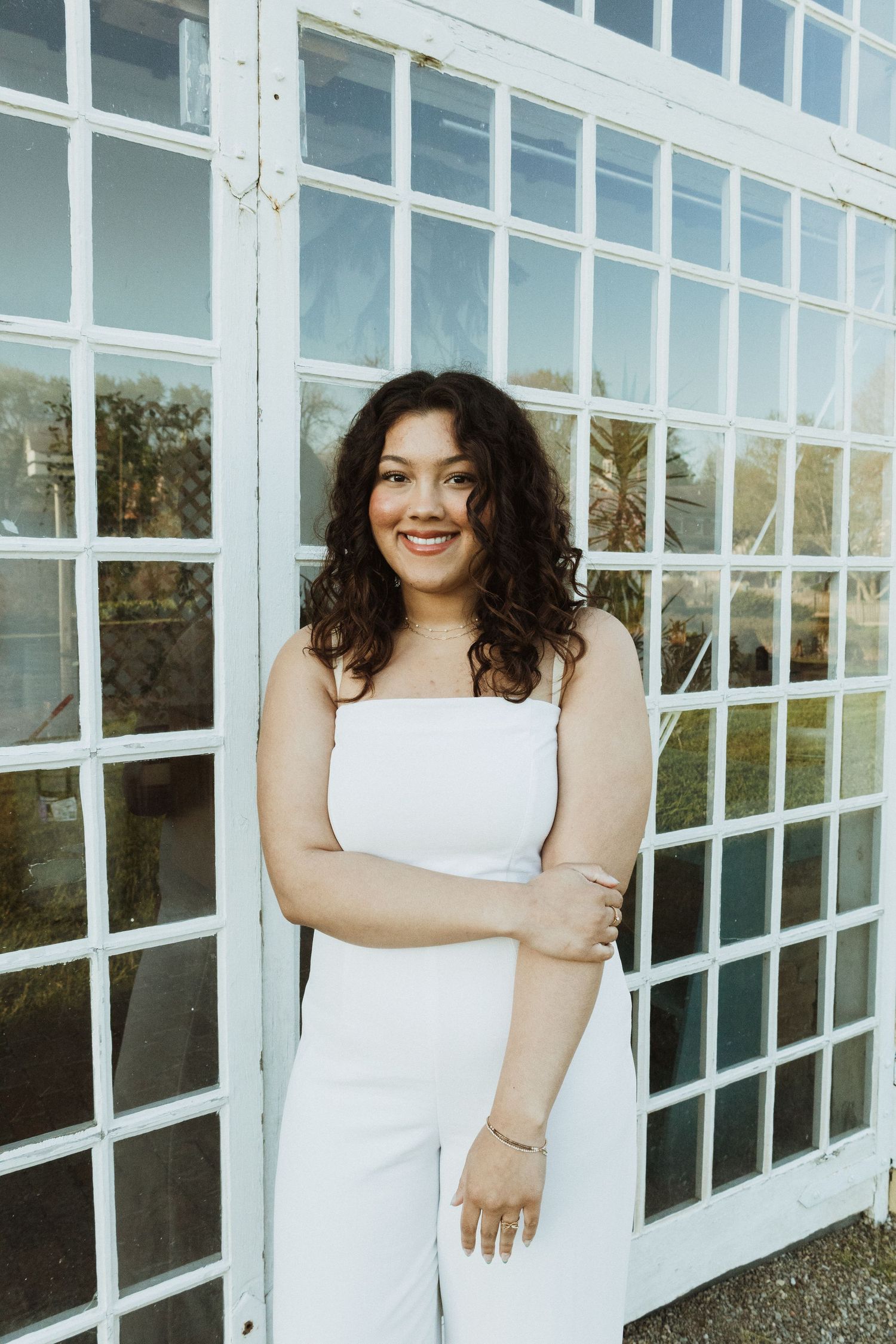 A woman in a white jumpsuit smiles brightly against a backdrop of white windowpanes.