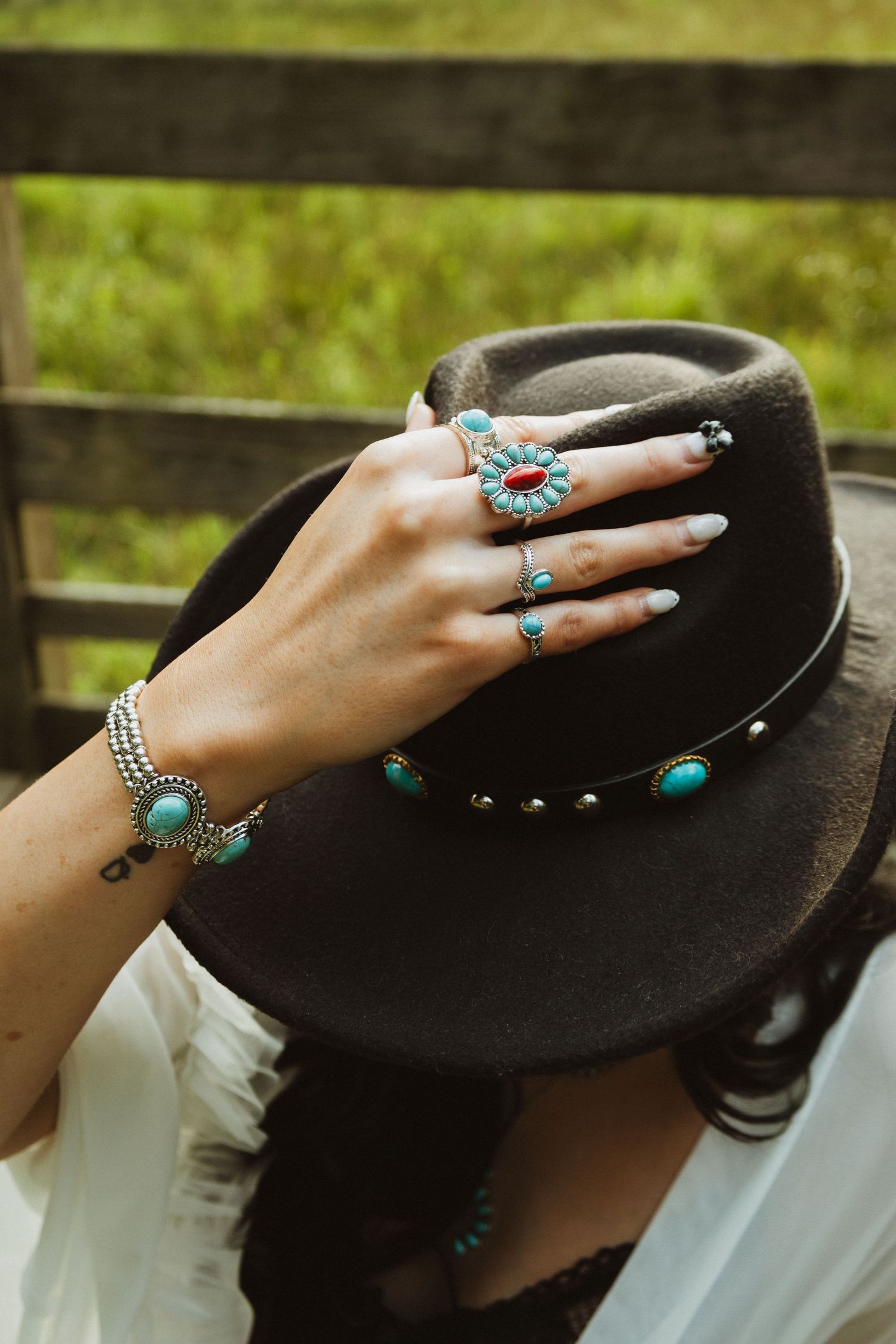 Hand adorned with turquoise rings rests on black wide-brimmed hat in artistic outdoor setting.