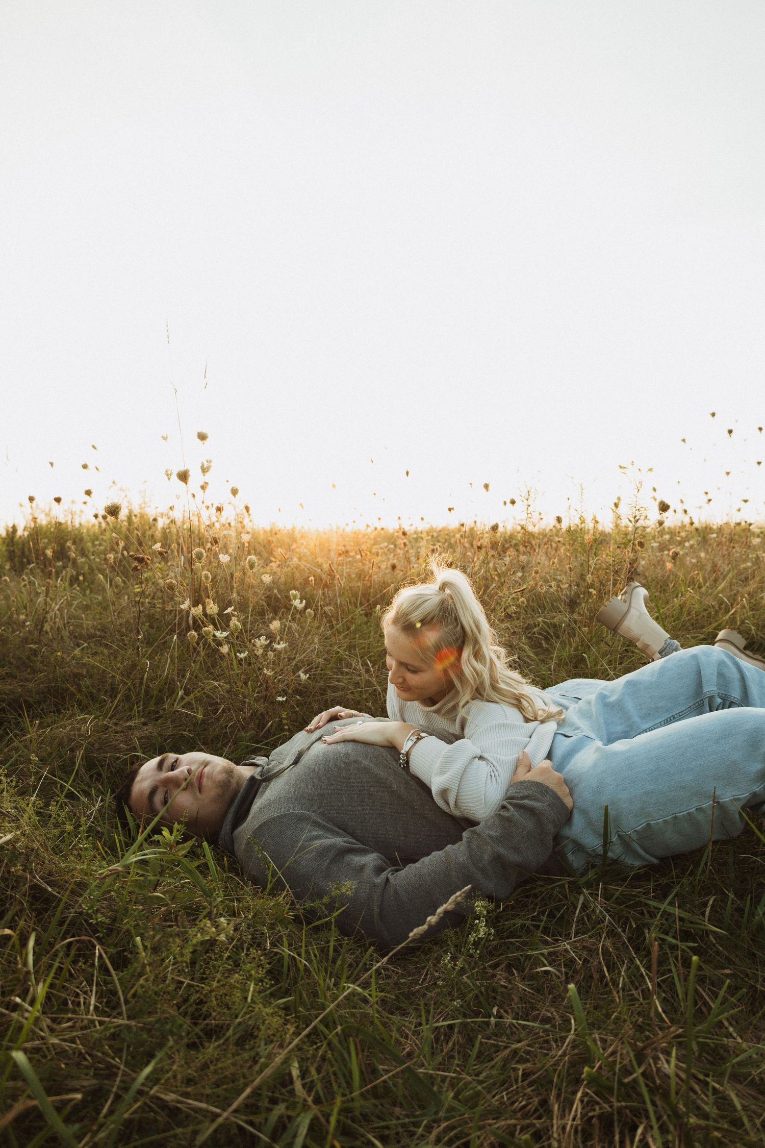 Two people lay together in tall grass during golden hour with wild flowers in the background.