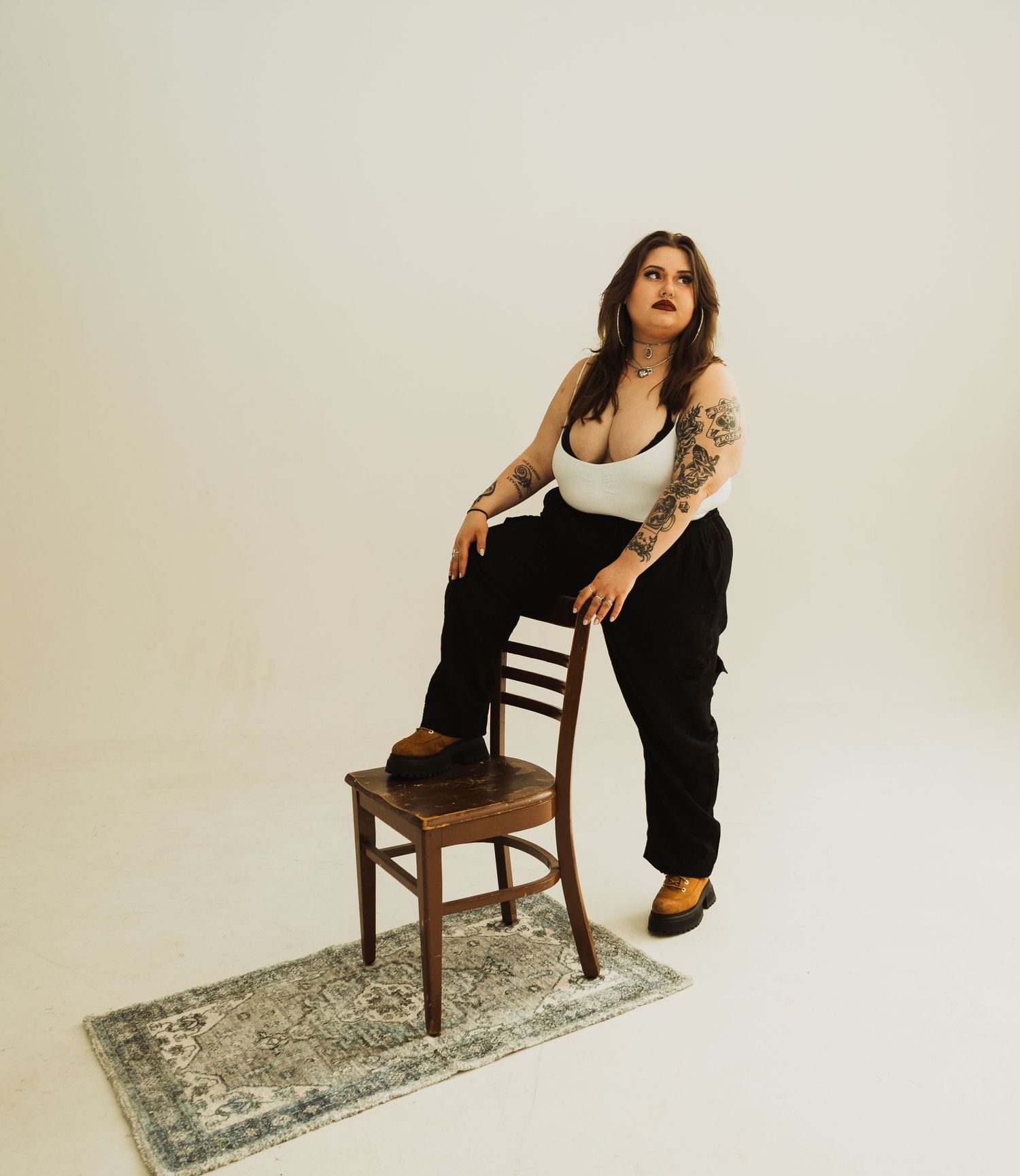 Figure in a white tank top and dark pants dramatically poses with tattoos on arms against a vintage wooden chair on a distressed rug.