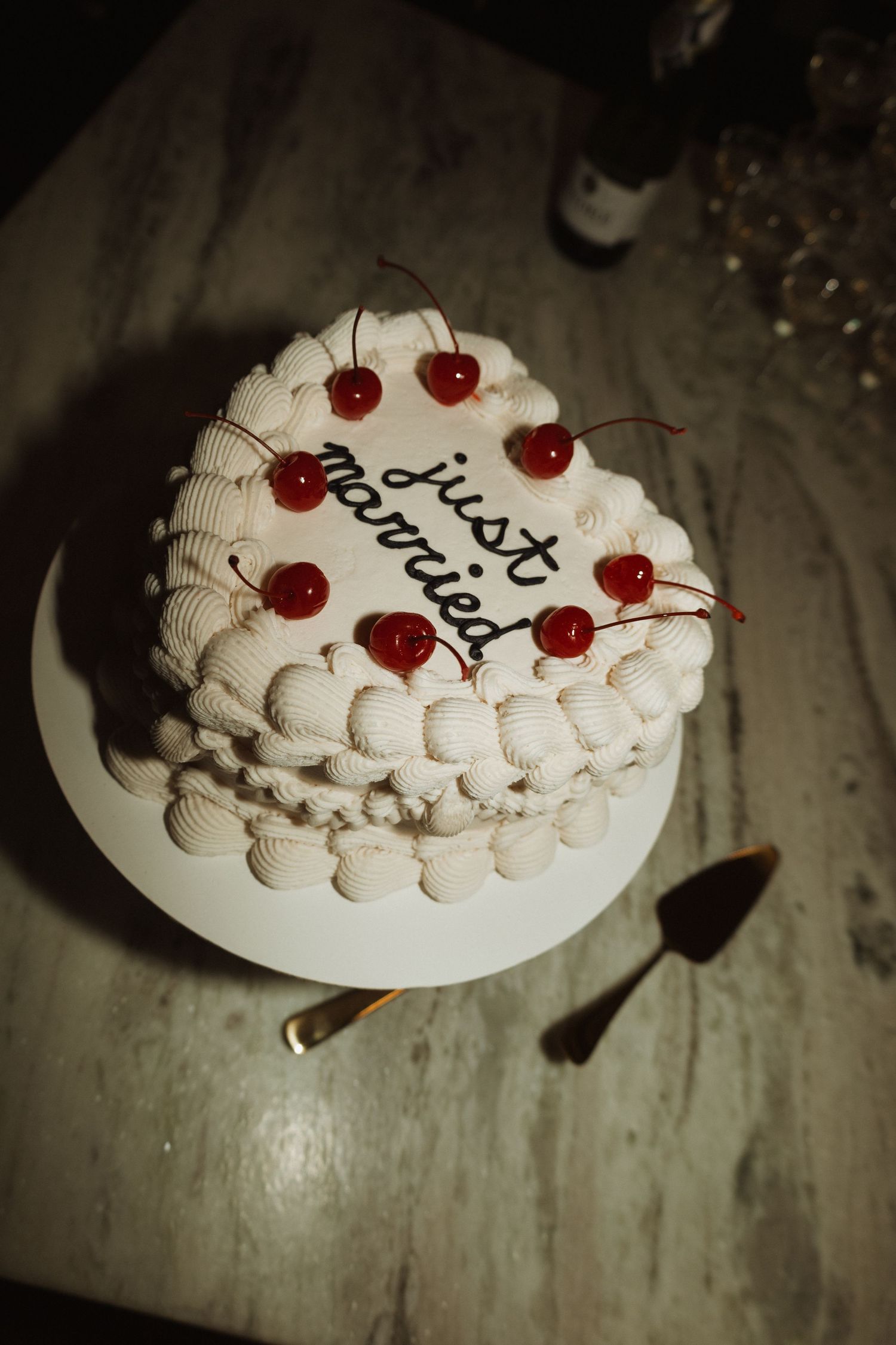 Heart-shaped white wedding cake with red cherries and black calligraphy in moody lighting.