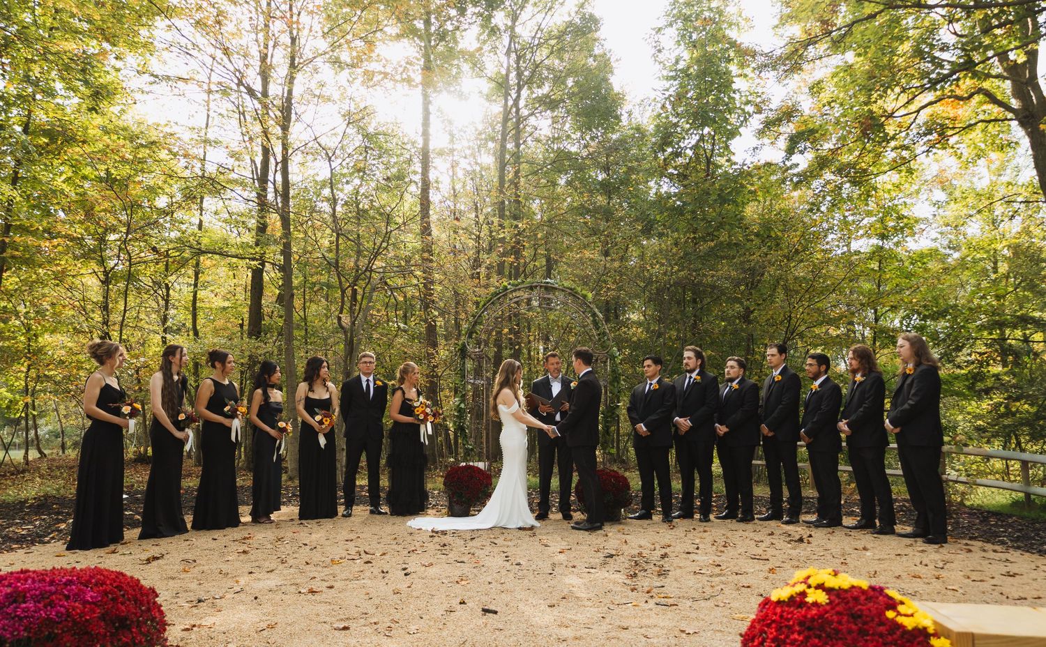 Wedding party lined up outdoors with fall trees and red flower petals on the ground.