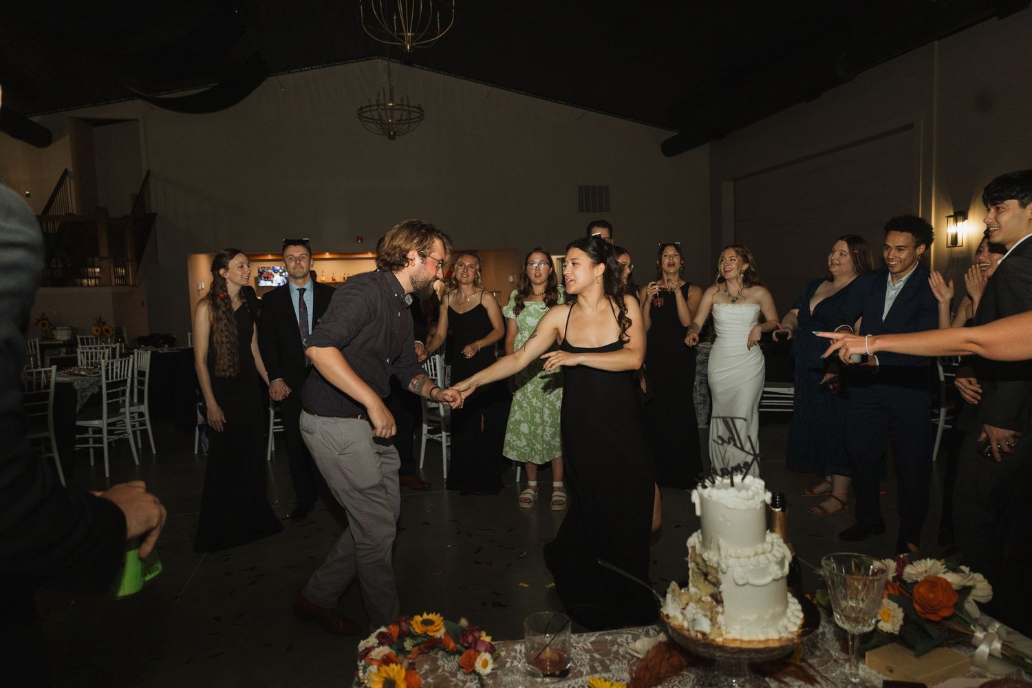 Wedding guests dance and socialize in an indoor venue with dark lighting and festive atmosphere.
