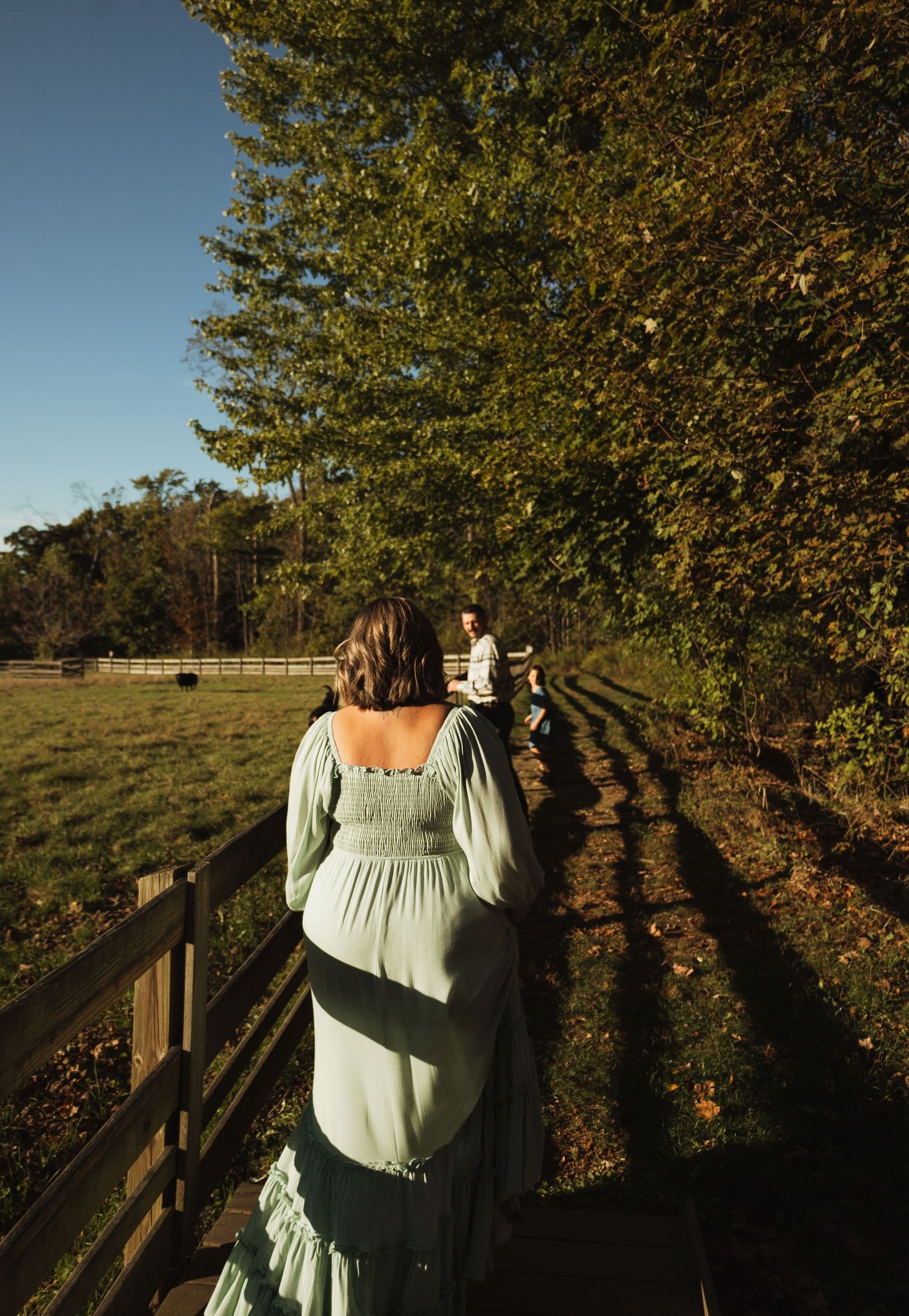 Person in light colored dress walking along wooden boardwalk with trees casting long shadows at sunset.