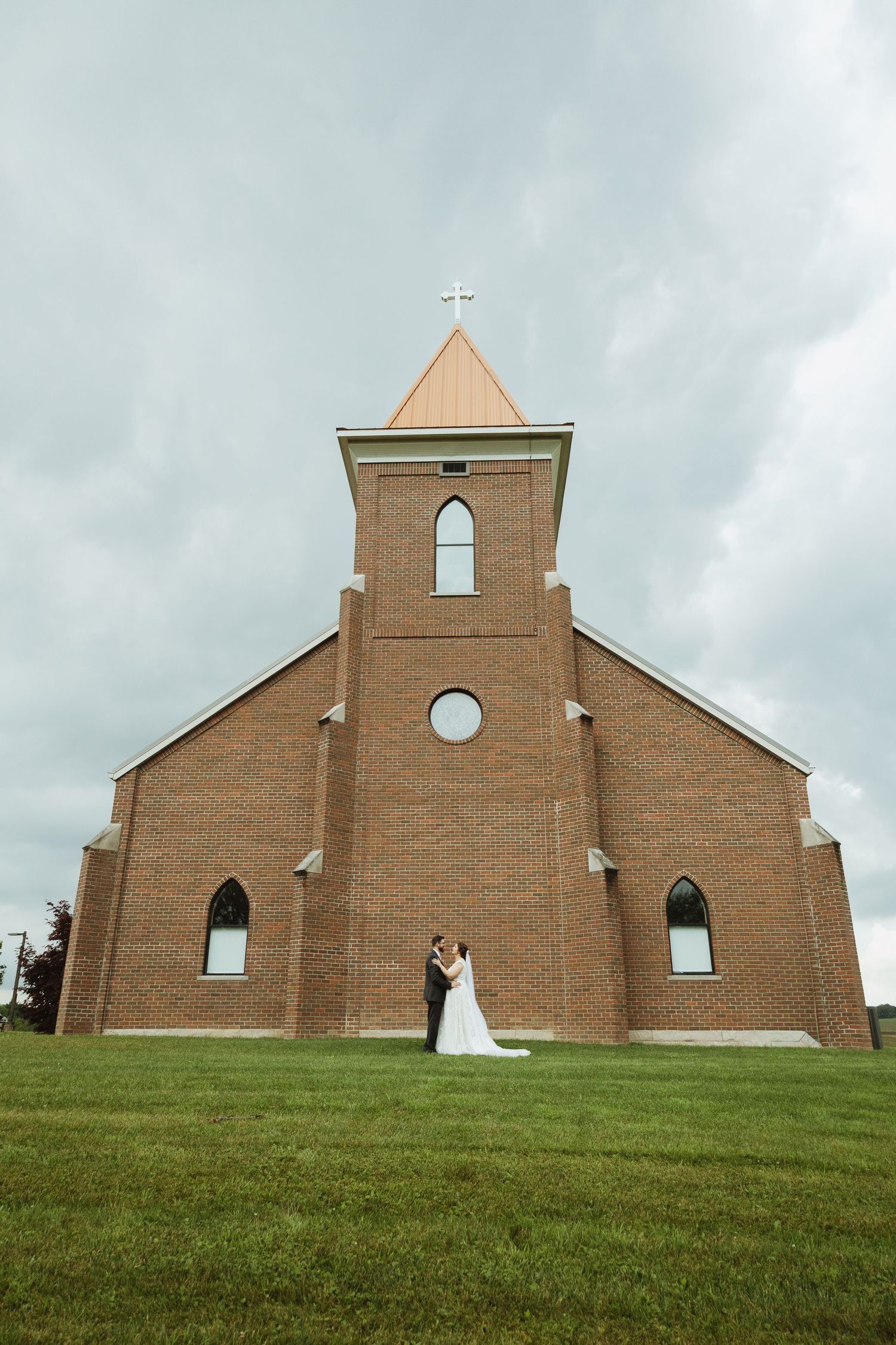 A traditional brick church with a tall steeple stands against cloudy skies as newlyweds pose on the lawn.