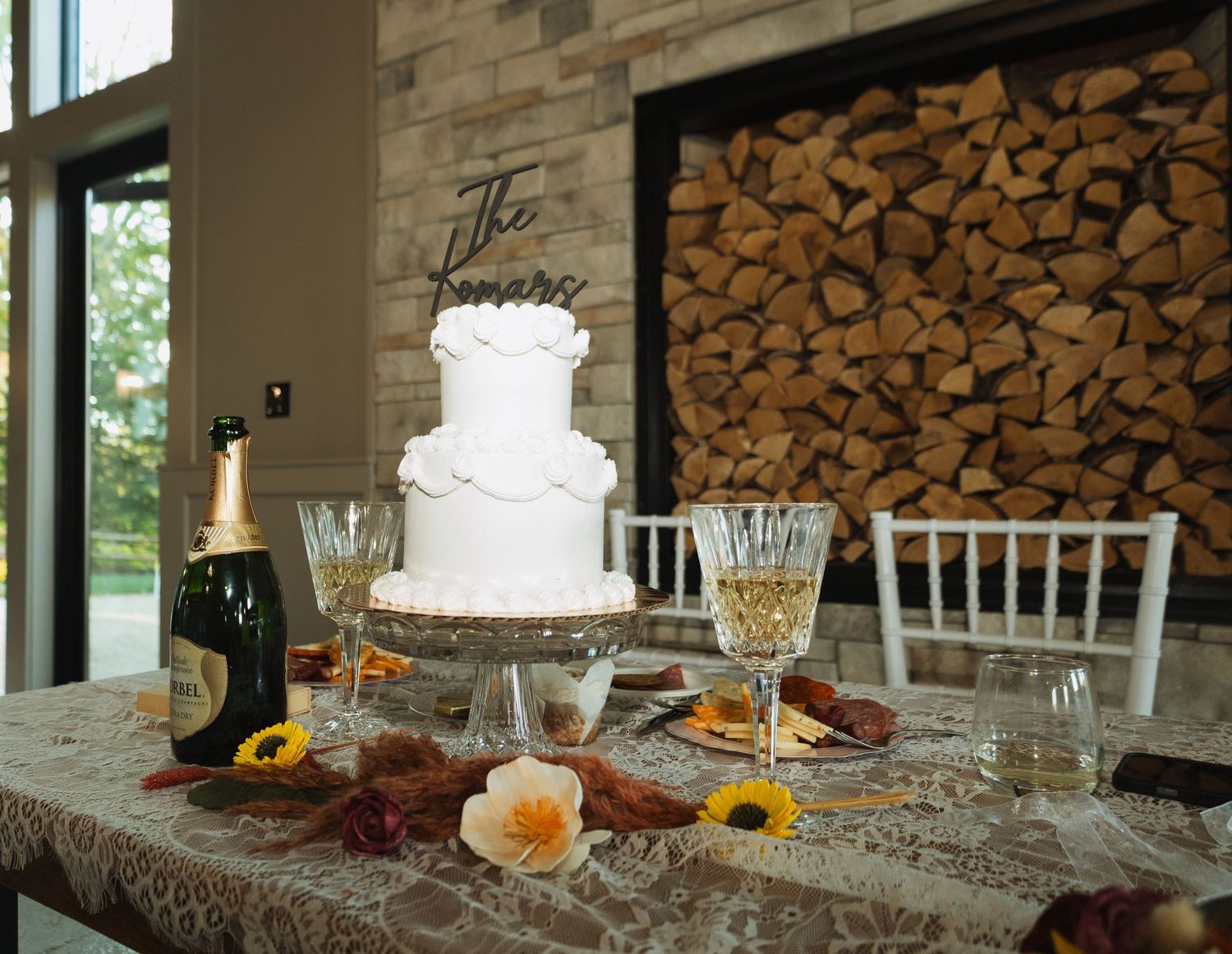 Elegant white wedding cake displayed on table with champagne glasses and rustic wood wall backdrop.