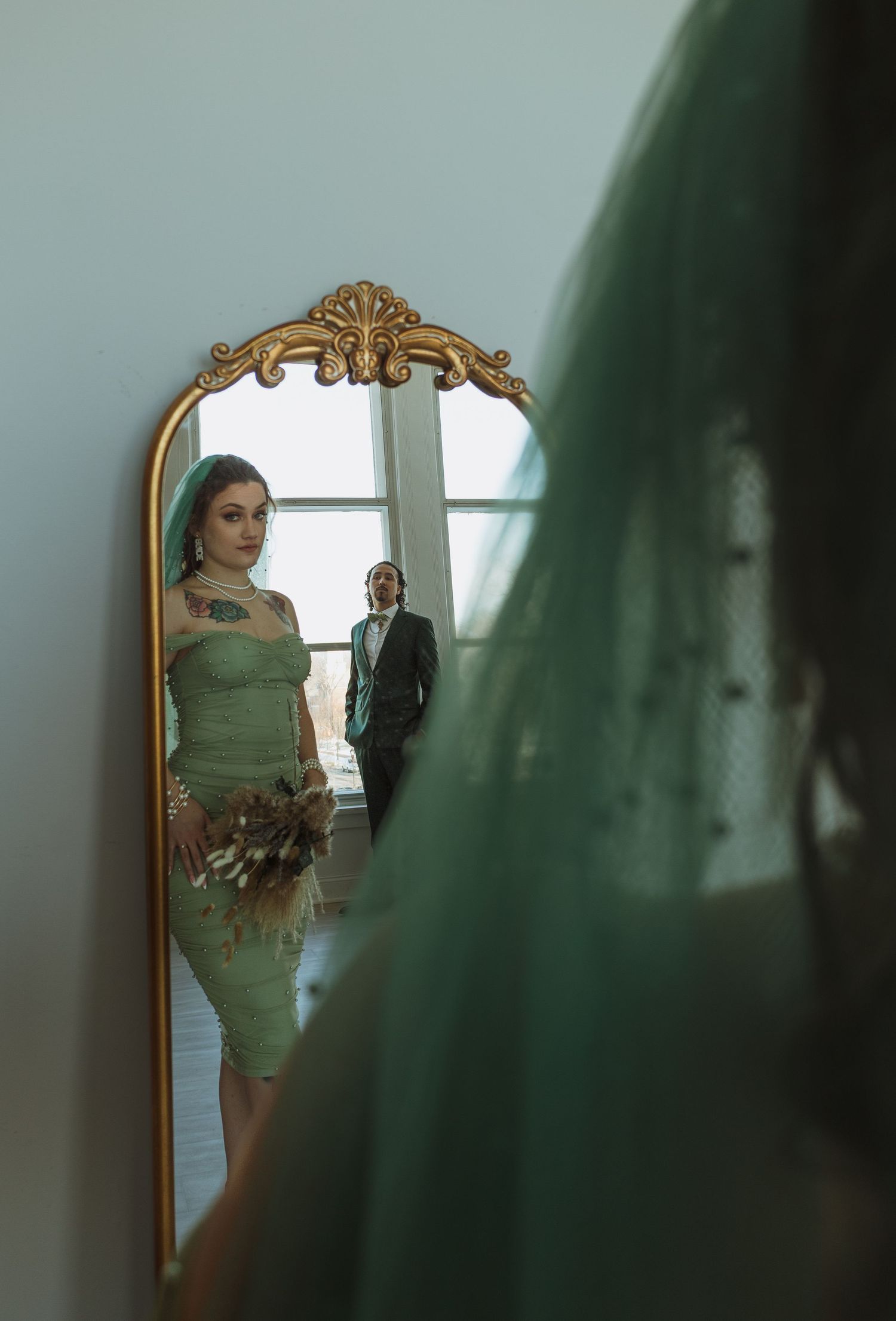 Bride in green lace dress and groom reflected in ornate gold mirror with teal veil foreground.