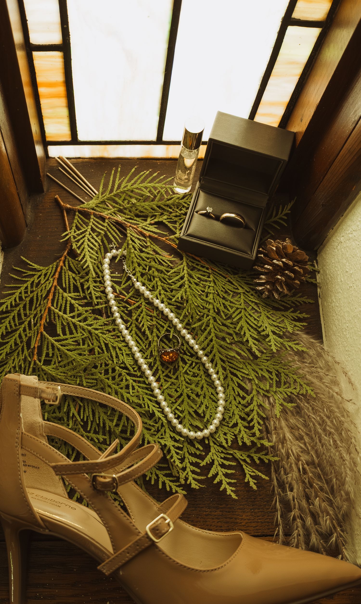 Flat lay of wedding accessories including pearl necklace, heels, ring box, perfume, and pine branches on wood.