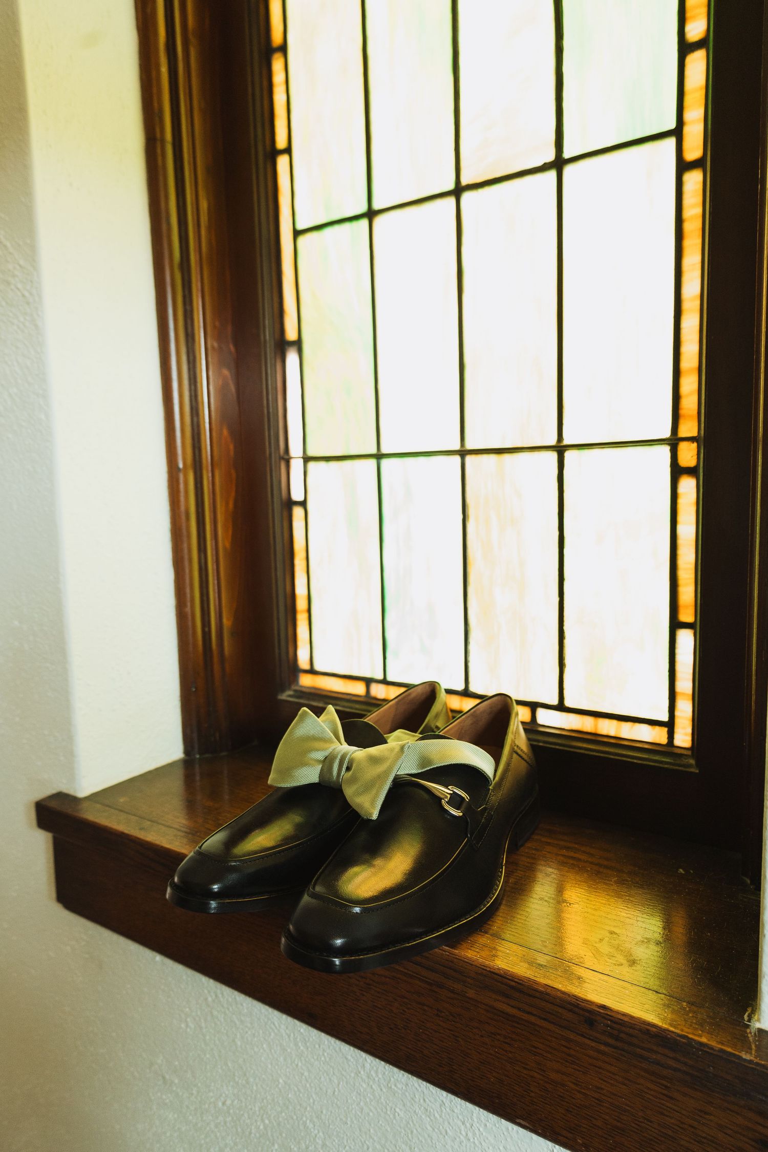 Black leather loafers with sage green bow tie displayed on wooden window sill with stained glass.