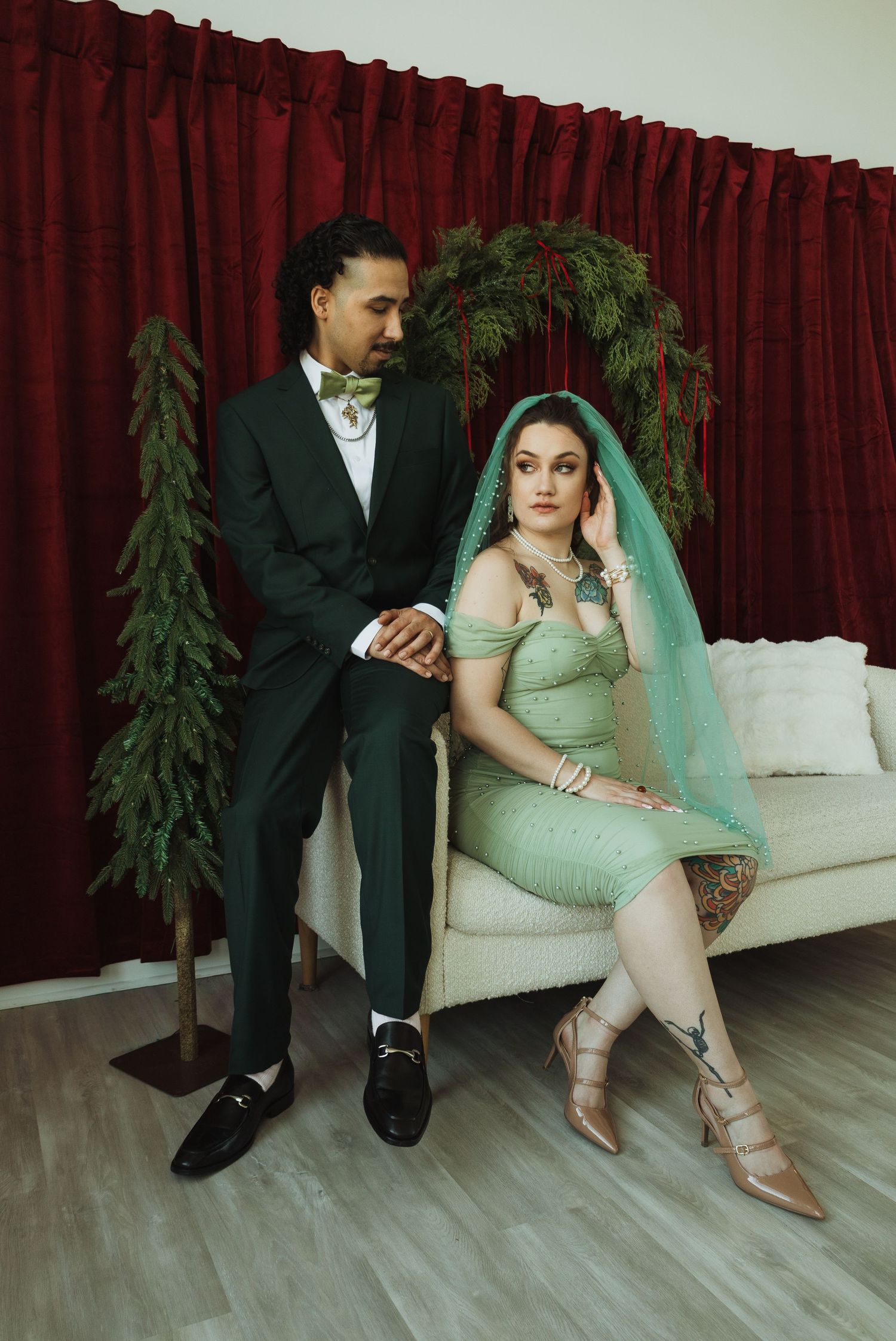 Couple in green wedding attire pose elegantly before red curtain backdrop with festive pine decor.