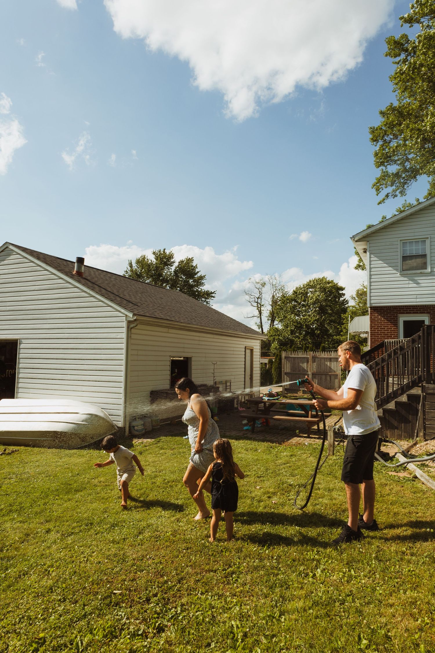 Family playing in backyard with garden hose on sunny day, with white house and brick building in background.