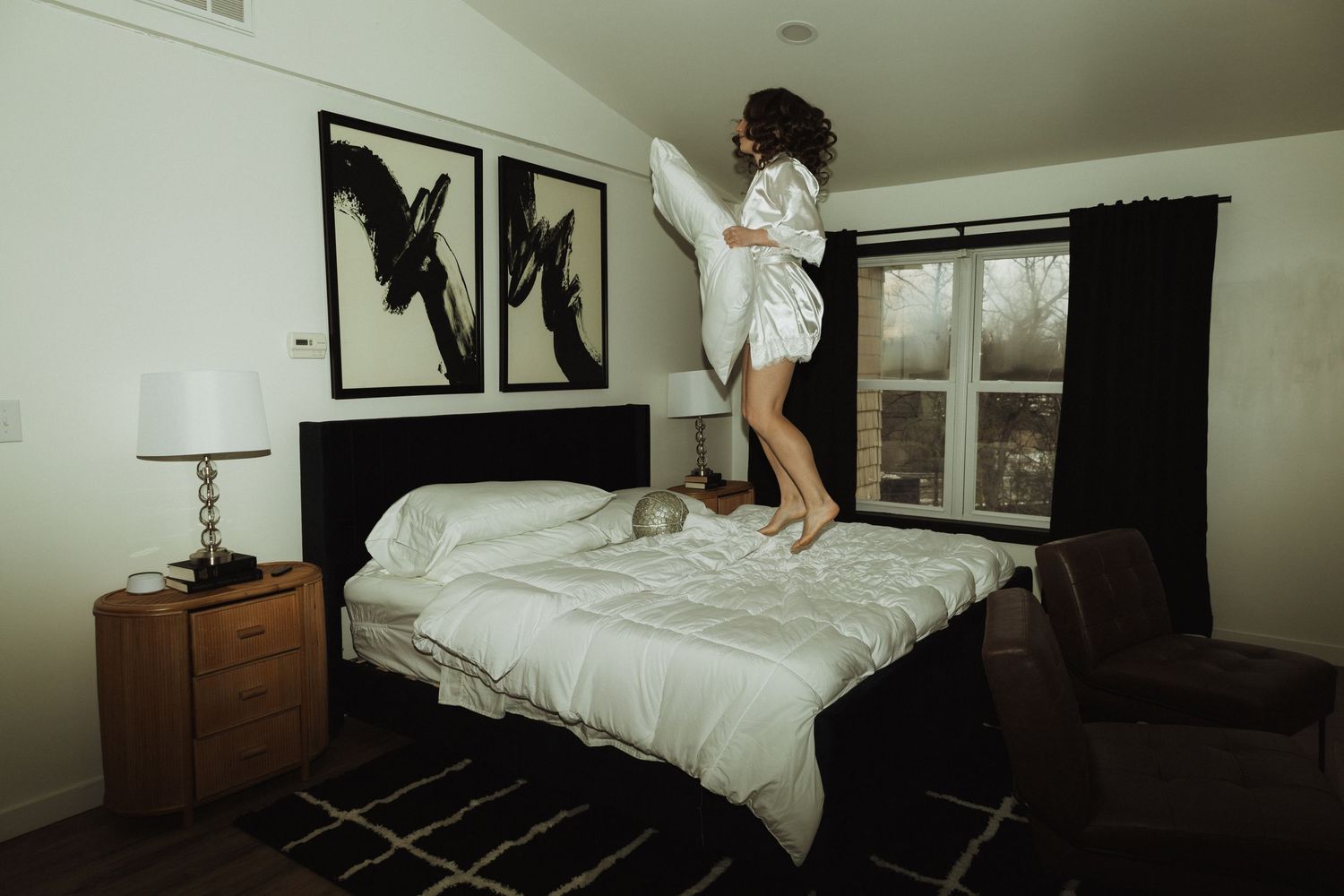 Person in white robe jumps playfully on bed with black artwork on wall.