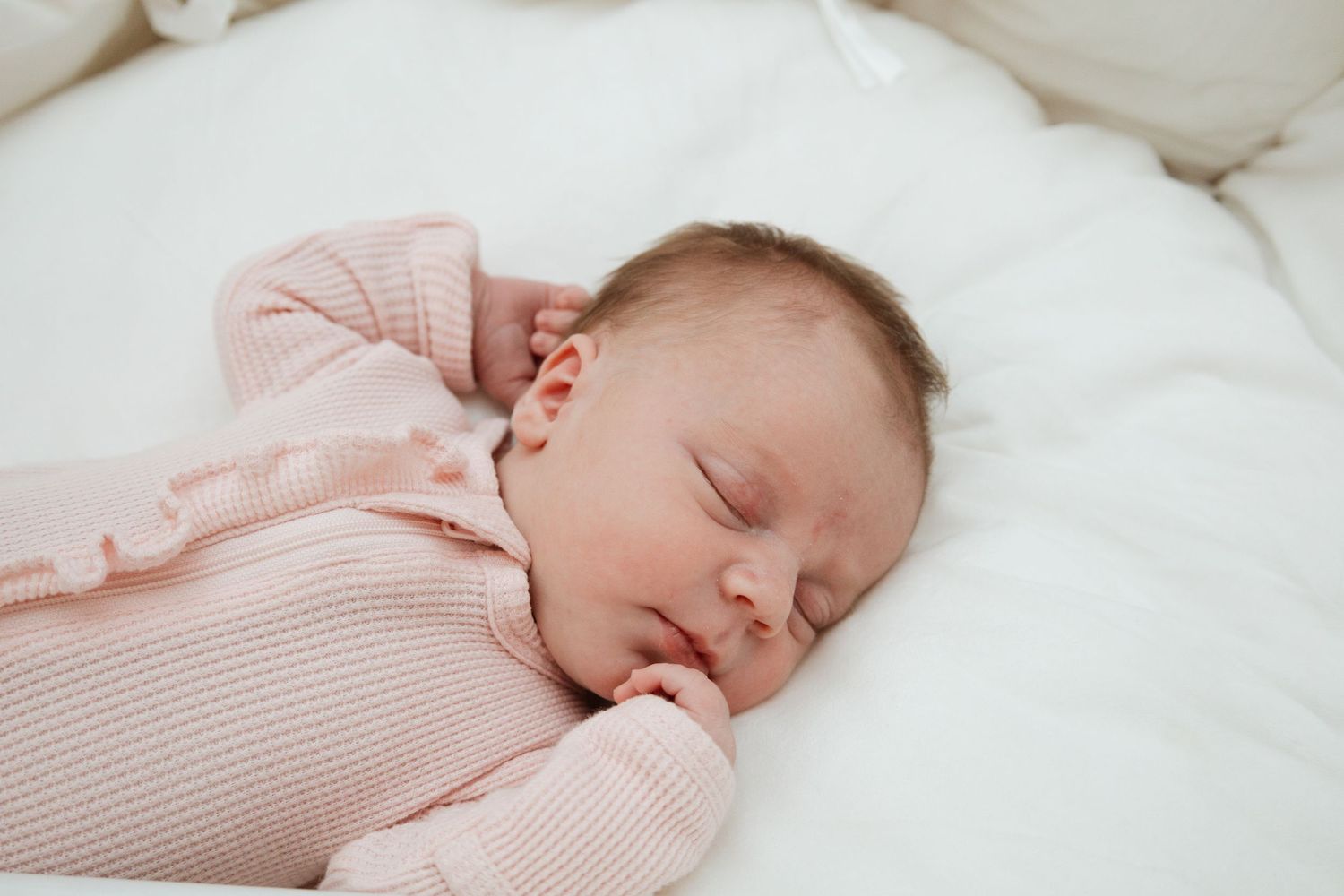 A sleeping baby wearing a soft pink knit sweater rests peacefully on white bedding.
