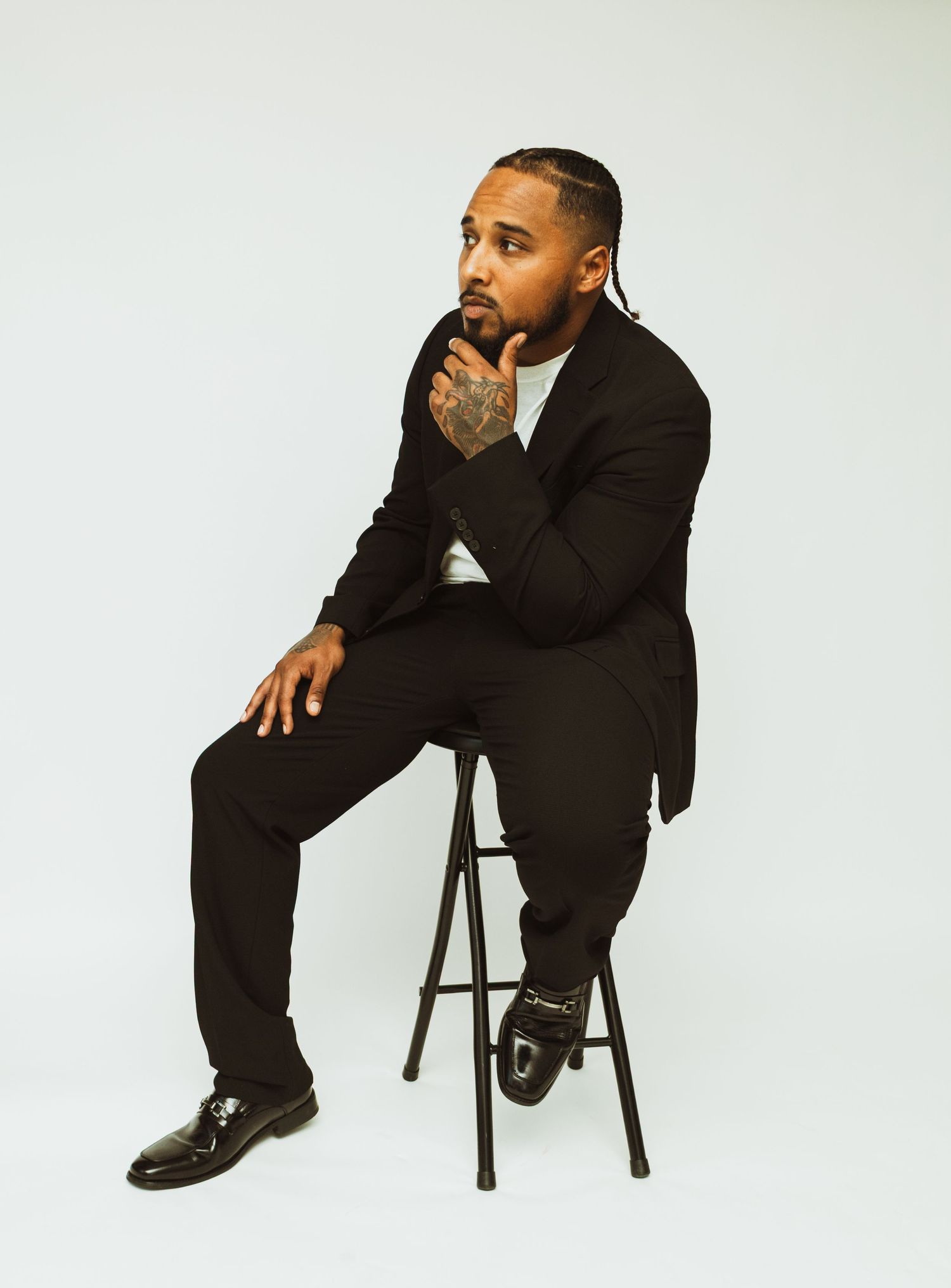 A man in a black suit sits thoughtfully on a barstool against a white backdrop.