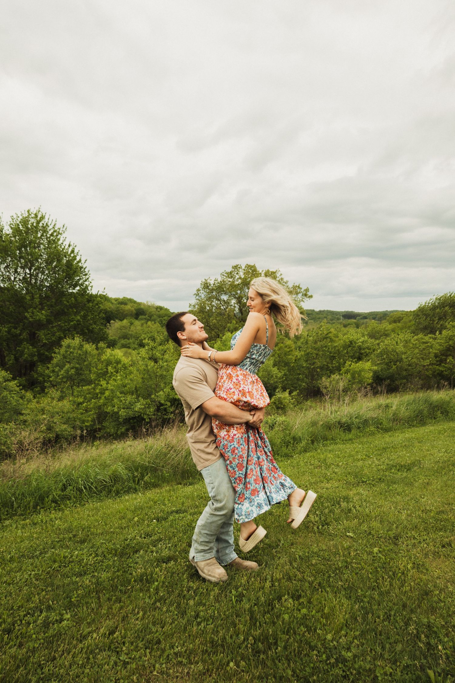 A couple embraces playfully while dancing on a grassy hillside with trees and cloudy skies in the background.