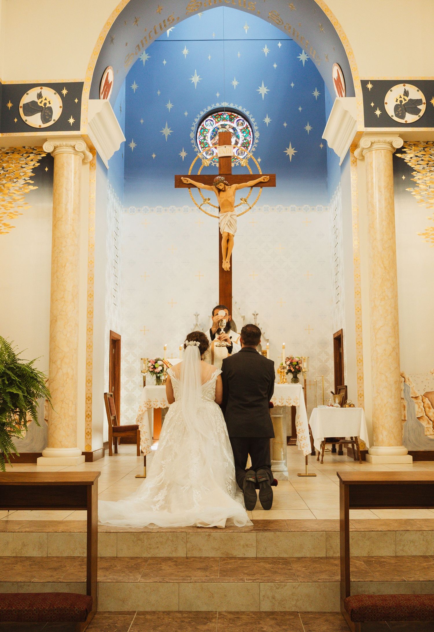 A wedding couple kneels at an altar in a chapel with white columns and a starry blue ceiling.