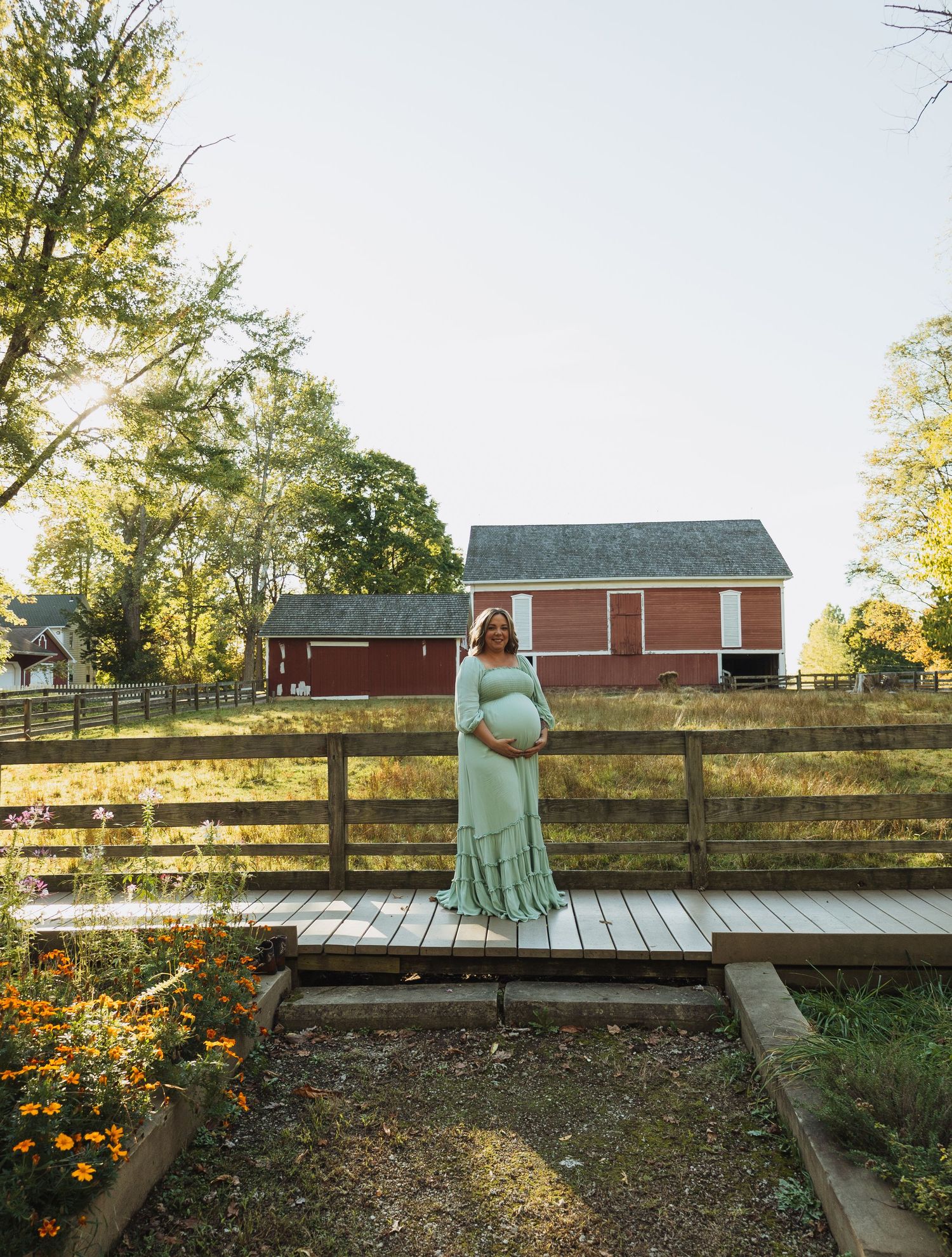 A rustic wooden deck and fence overlooks a garden with orange marigolds at sunset near a red barn.