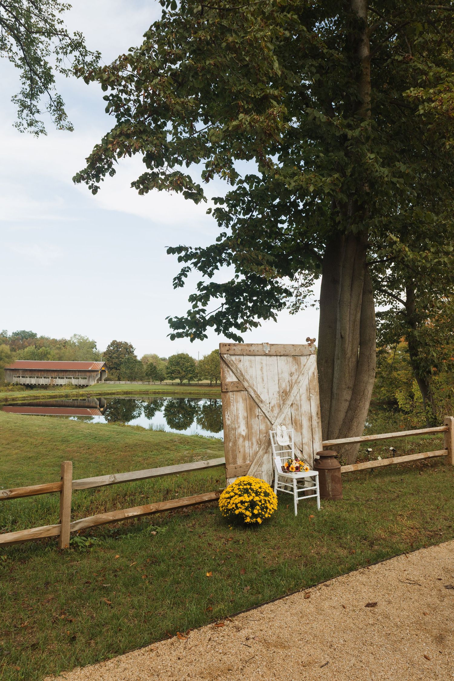 Rustic wedding decor featuring a vintage door and chair against a wooden fence with yellow mums and pond view.