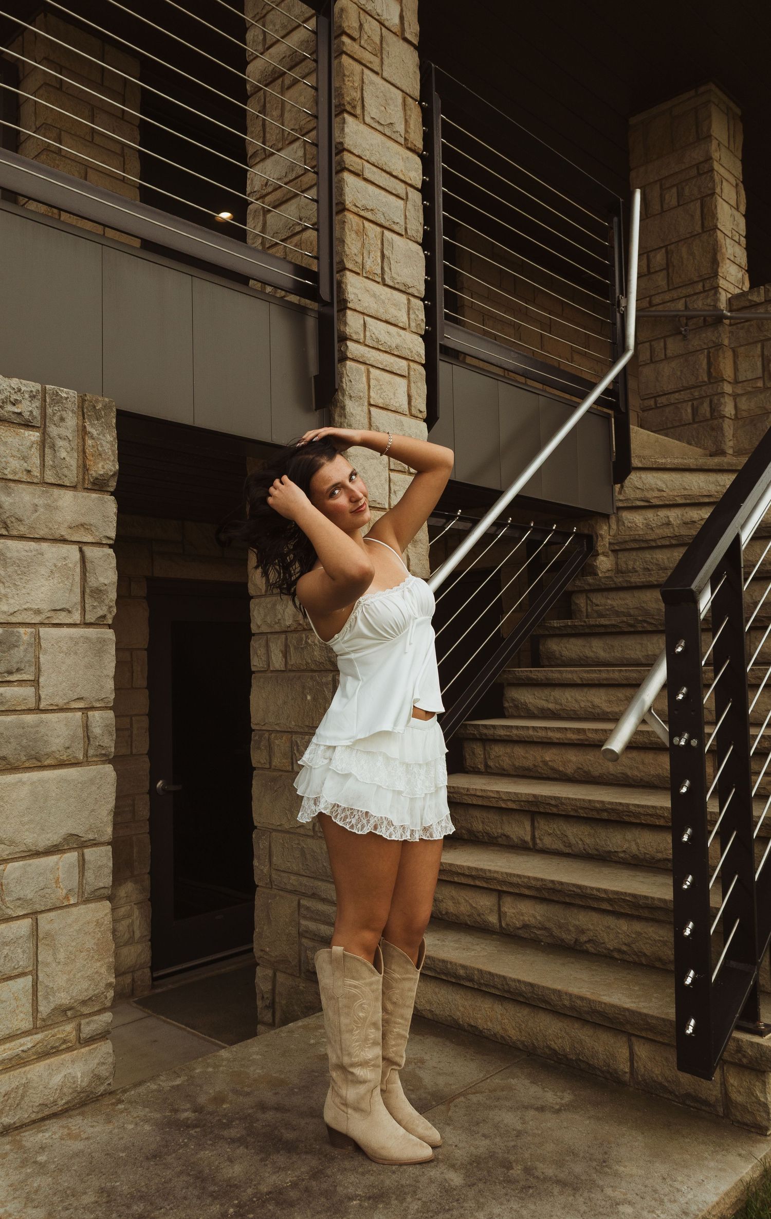Young woman in white ruffled mini skirt, crop top, and cowboy boots posing by stone building staircase.