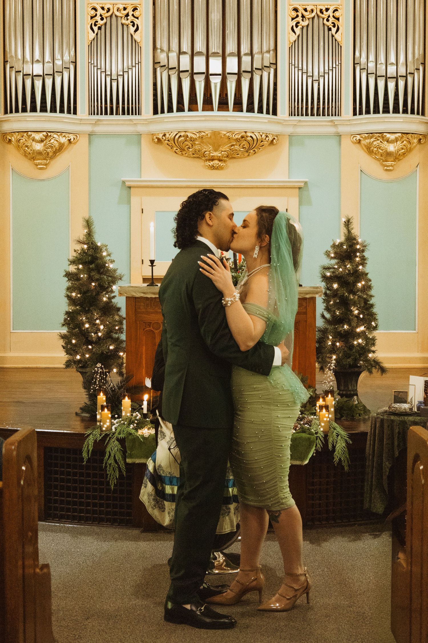 Bride and groom share first kiss at church altar with pipe organ, Christmas trees, and candlelight.