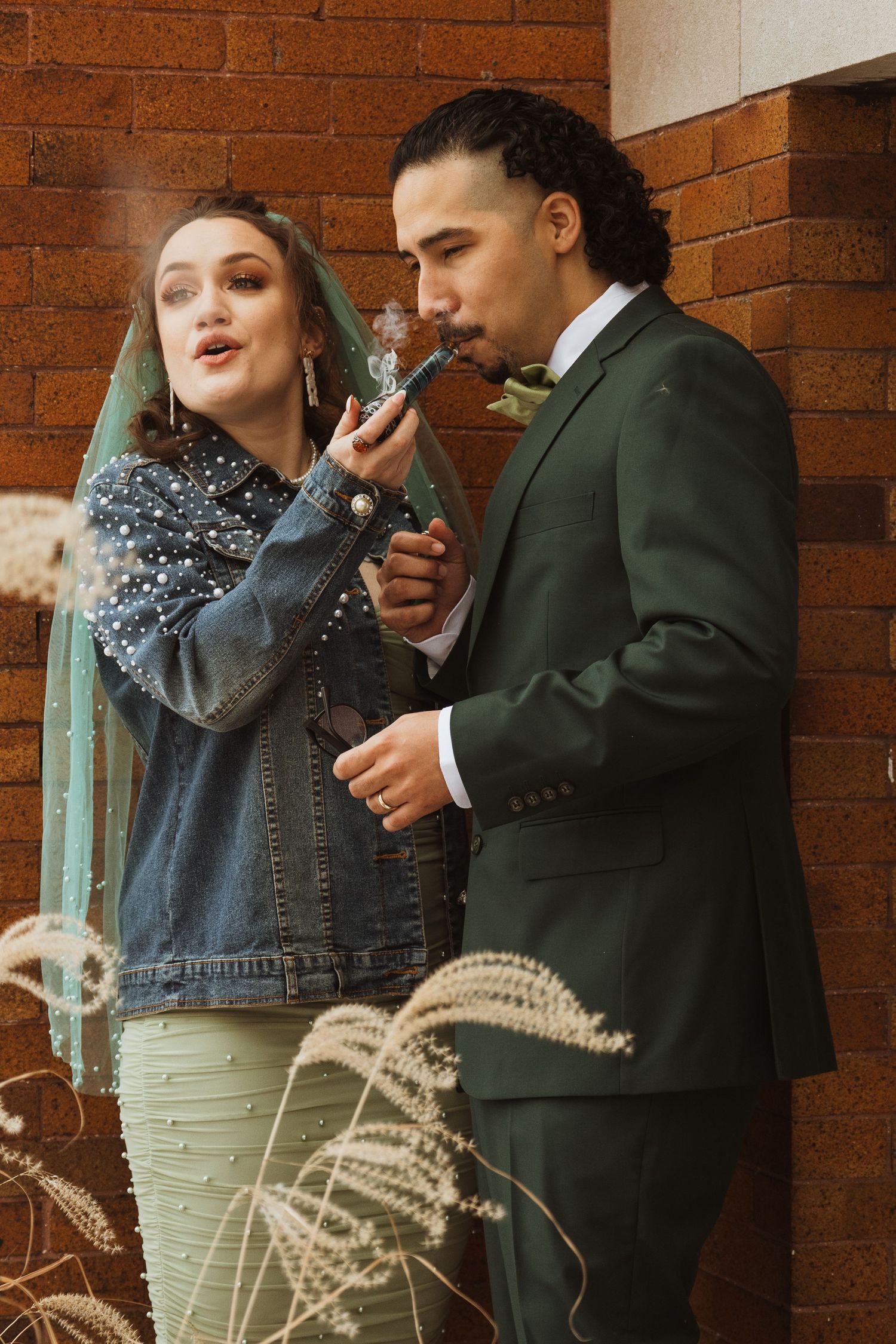 Bride in denim jacket and veil lighting groom's cigarette against rustic brick wall outdoors.