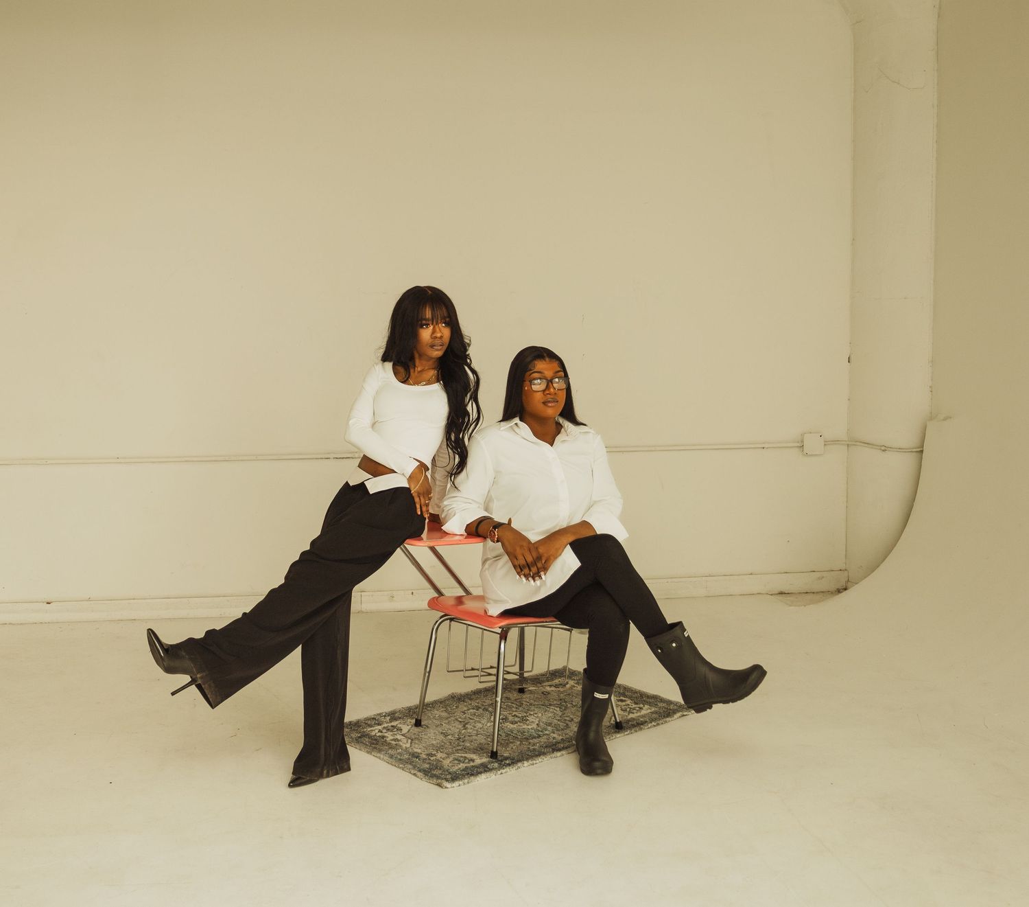Two people in coordinated white and black outfits posing on metallic chair in studio.