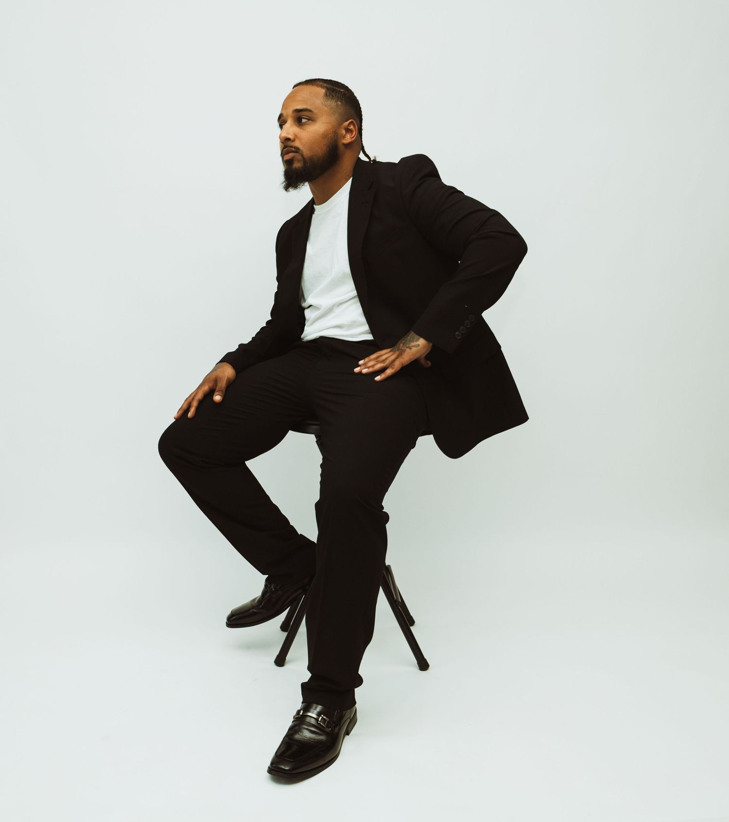 A person in a black suit and white shirt poses confidently on a stool against a white studio backdrop.