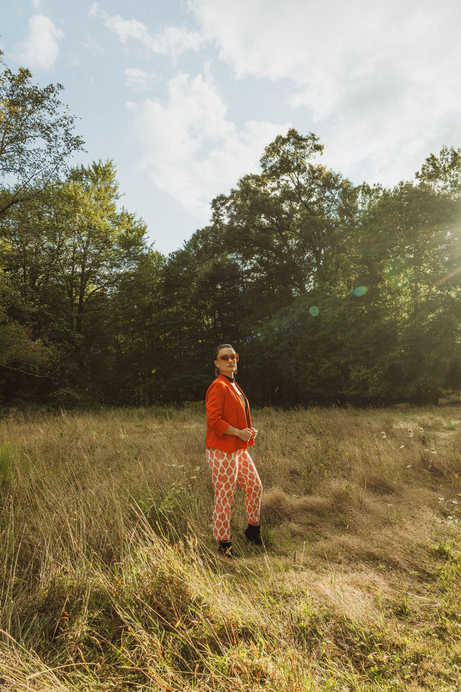 Full-length shot of trendy outfit featuring orange blazer and patterned pants in a rustic field setting.