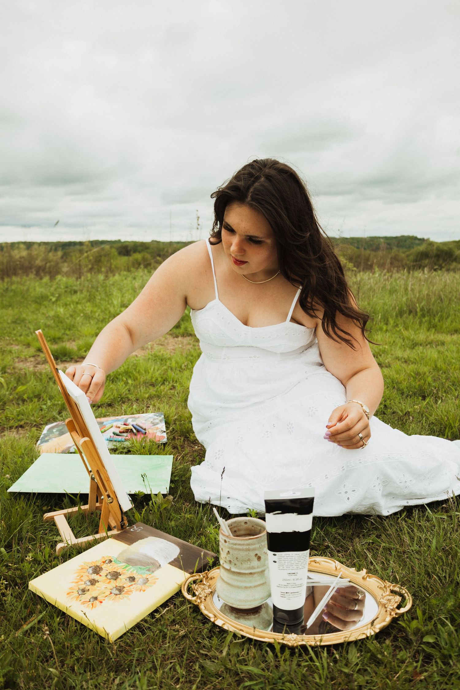 Artist in white dress painting on canvas while sitting in grassy meadow with art supplies and drinks on ornate tray.