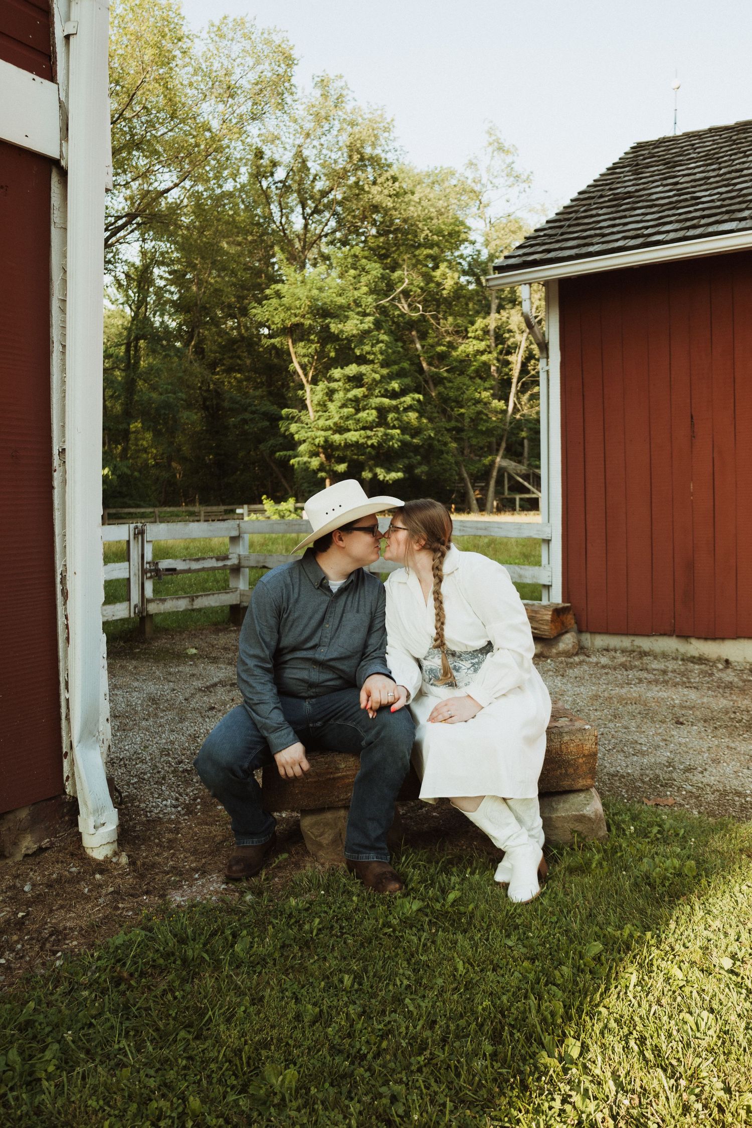 A romantic couple rests together on steps of a red barn during golden hour.