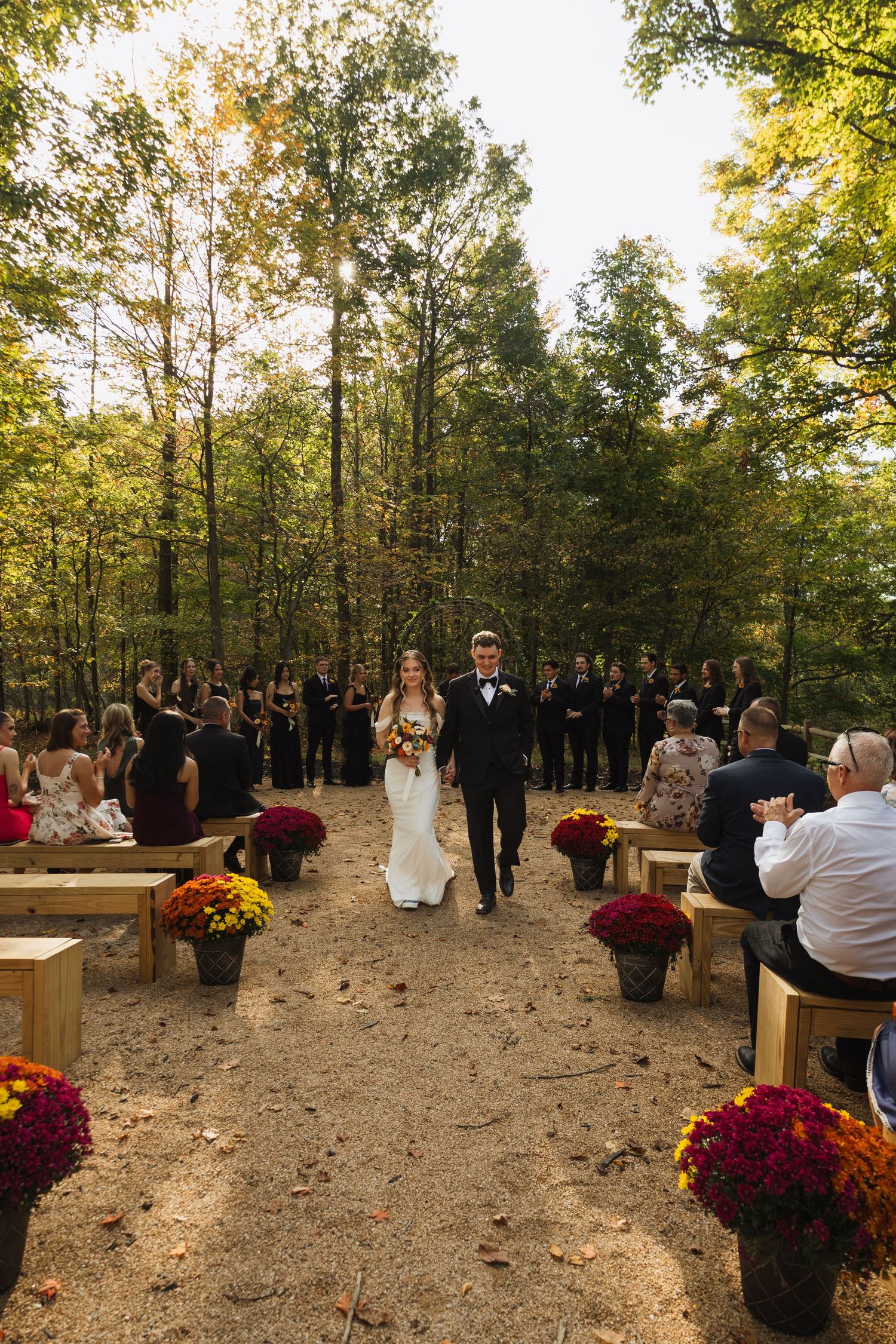 Outdoor forest wedding ceremony with wooden benches and fall flowers in natural setting.